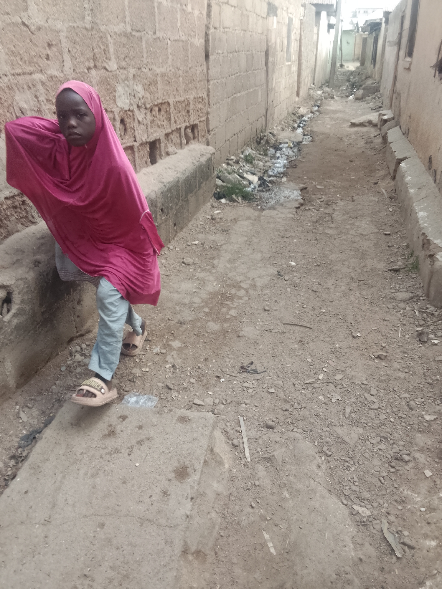 A young person wearing a pink headscarf, light blue pants, and sandals stands in an alleyway in Pantame, Nigeria. The alley is narrow, with brick walls on either side, and debris and trash scattered along the edges. The ground is a mix of dirt and concrete.