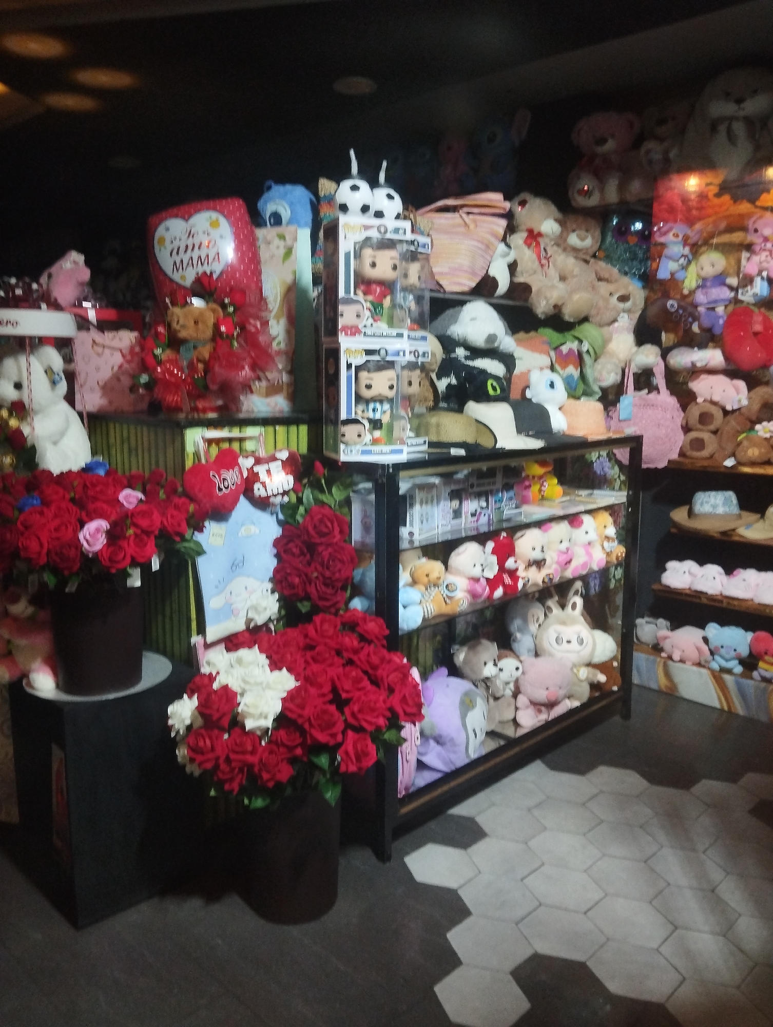 The image depicts a retail space filled with a variety of plush toys, figurines, and floral arrangements. The location is identified as El Valle del Espíritu Santo, Venezuela.
The foreground features two large floral arrangements, one with red roses and a second with red and white roses, both in dark brown cylindrical vases. These are positioned on dark, square display stands. Behind the floral arrangements are shelves and displays stocked with stuffed animals, including teddy bears, and other plush characters. Several "Funko Pop" figurines in boxes are visible on a shelf.
In the background, more plush toys are displayed, including various types of stuffed animals and dolls. A balloon with the words "te amo mama" is visible among the items. The room is dimly lit, and the floor is covered with grey, hexagon-shaped tiles. There is no presence of people in this image.