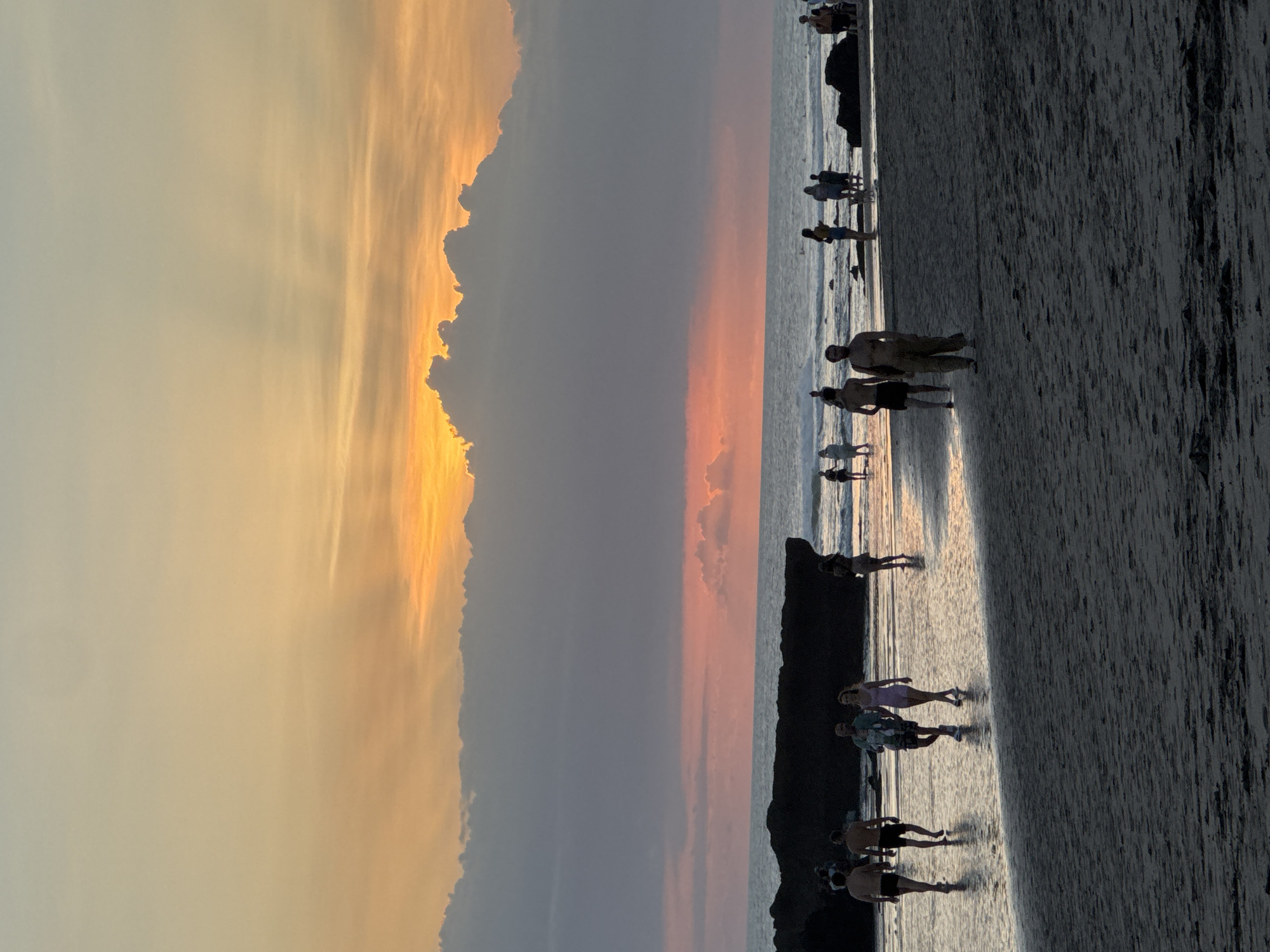 Captured in North Kuta, Indonesia, the image depicts a beach scene at sunset. Silhouetted figures of individuals, mostly in casual attire, dot the shoreline, some wading in the shallows, while others are walking along the wet sand. The sky exhibits a gradient of colors, from the pale yellow near the horizon to a deeper orange and gray as it rises, reflecting the sunset's glow onto the sea. Dark, rocky formations are visible along the beach.