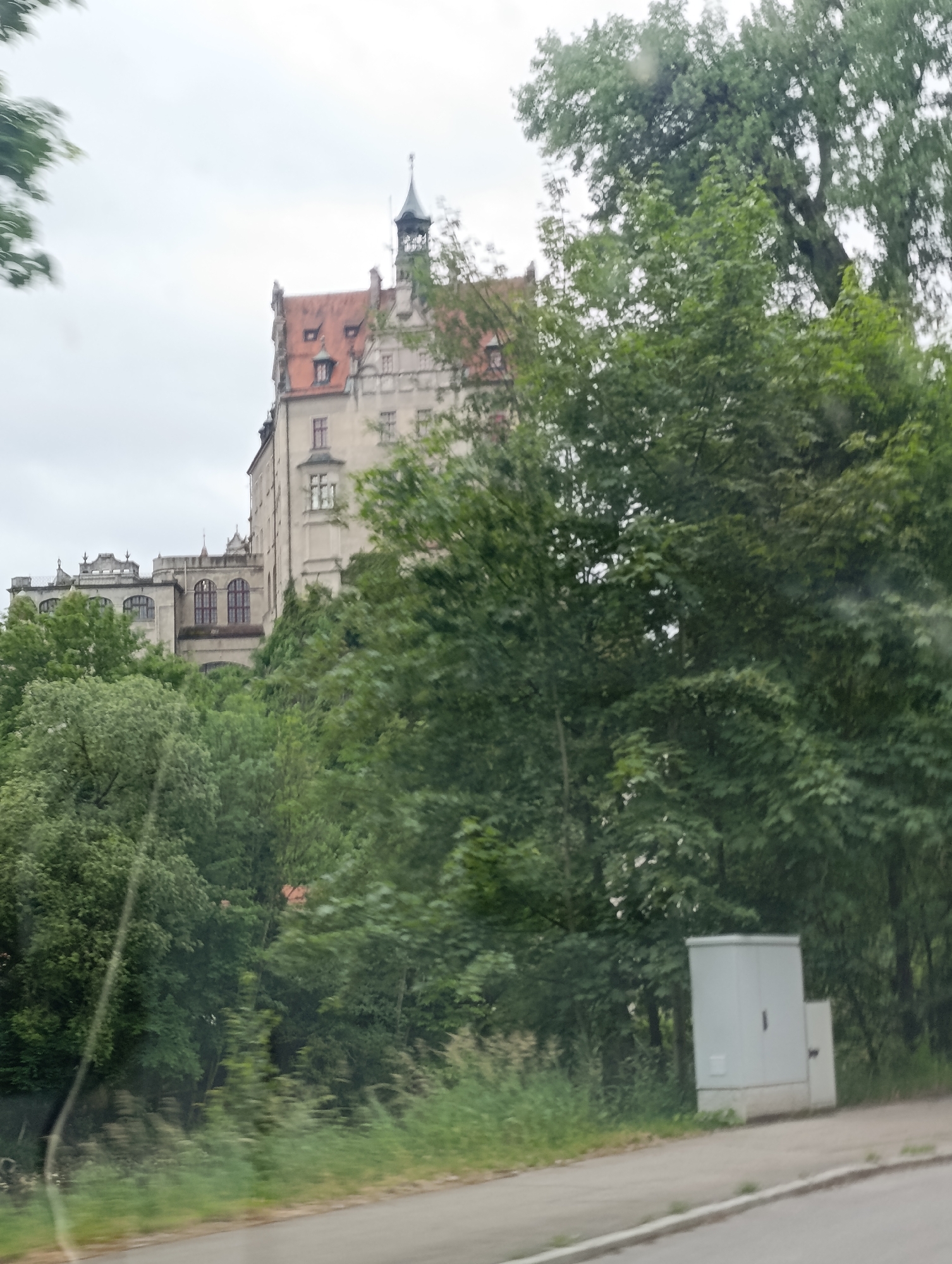 The image depicts a large, historic building, likely a castle or palace, situated atop a hill. The architecture features a reddish-orange tiled roof, pale stone walls, and multiple windows, some of which are arched. A tall, narrow spire with a decorative finial crowns a section of the building, adding to its grandeur. The castle is partially obscured by lush green trees and foliage, suggesting a natural or park-like setting. In the foreground, a paved road or path runs alongside the scene, with a modern, grey electrical cabinet or utility box placed on a concrete base near the edge of the path. The overall impression is that of an old-world structure integrated with a natural landscape.
The time of day appears to be overcast, with a diffused light suggesting either early morning or a cloudy afternoon. The weather seems to be mild, as evidenced by the abundant greenery and the absence of any harsh shadows or bright sunlight. The location is identified as Sigmaringen, Germany. No people are visible in the image, and there is no discernible text. The scene evokes a sense of historical significance and natural beauty.