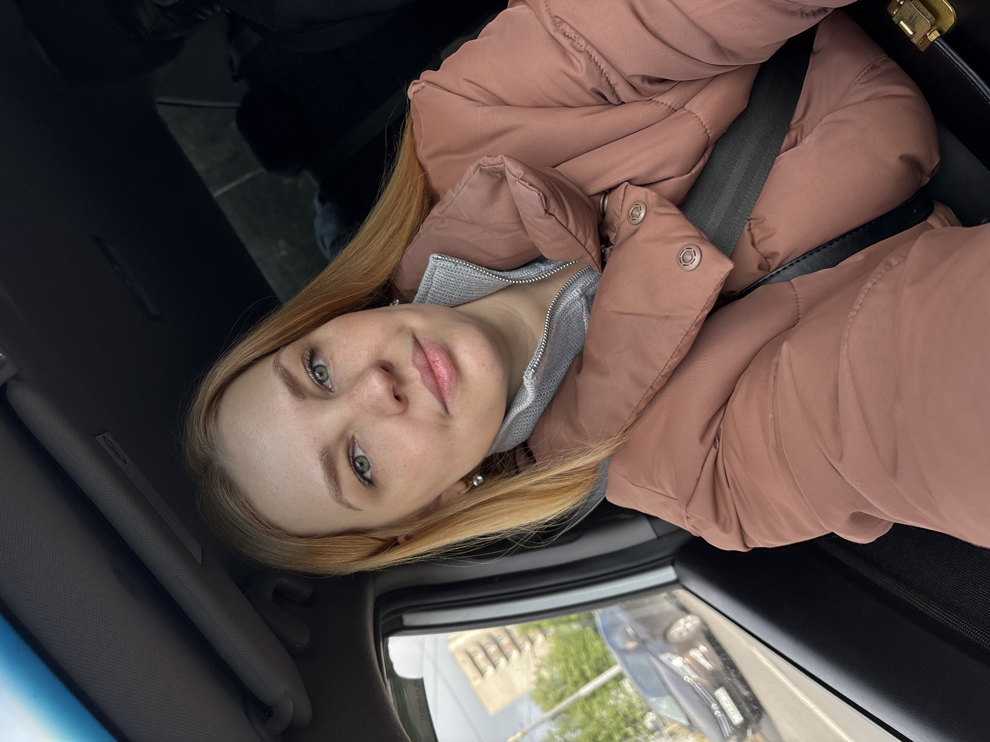 A young woman with fair skin and light blonde hair is wearing a pinkish-brown puffy jacket and a grey zippered shirt. She is inside a car, wearing a seatbelt. Outside the car window, a building and trees are visible, as well as the reflection of another vehicle. The location may be Fedorino, Russia.