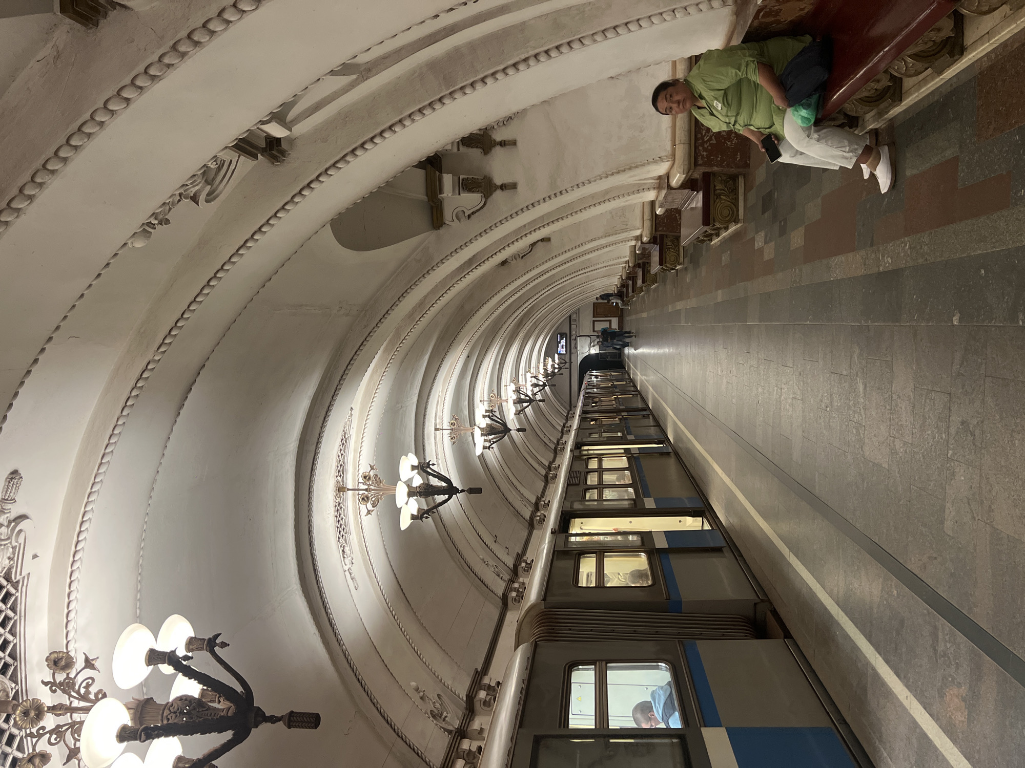 The image depicts a subway station. The architecture features arched ceilings adorned with decorative moldings and ornate chandeliers. A train is visible along the platform, partially obscuring the view further down the tunnel. A person sits on a bench alongside the platform. The subway station is in the city of Unknown, Unknown.