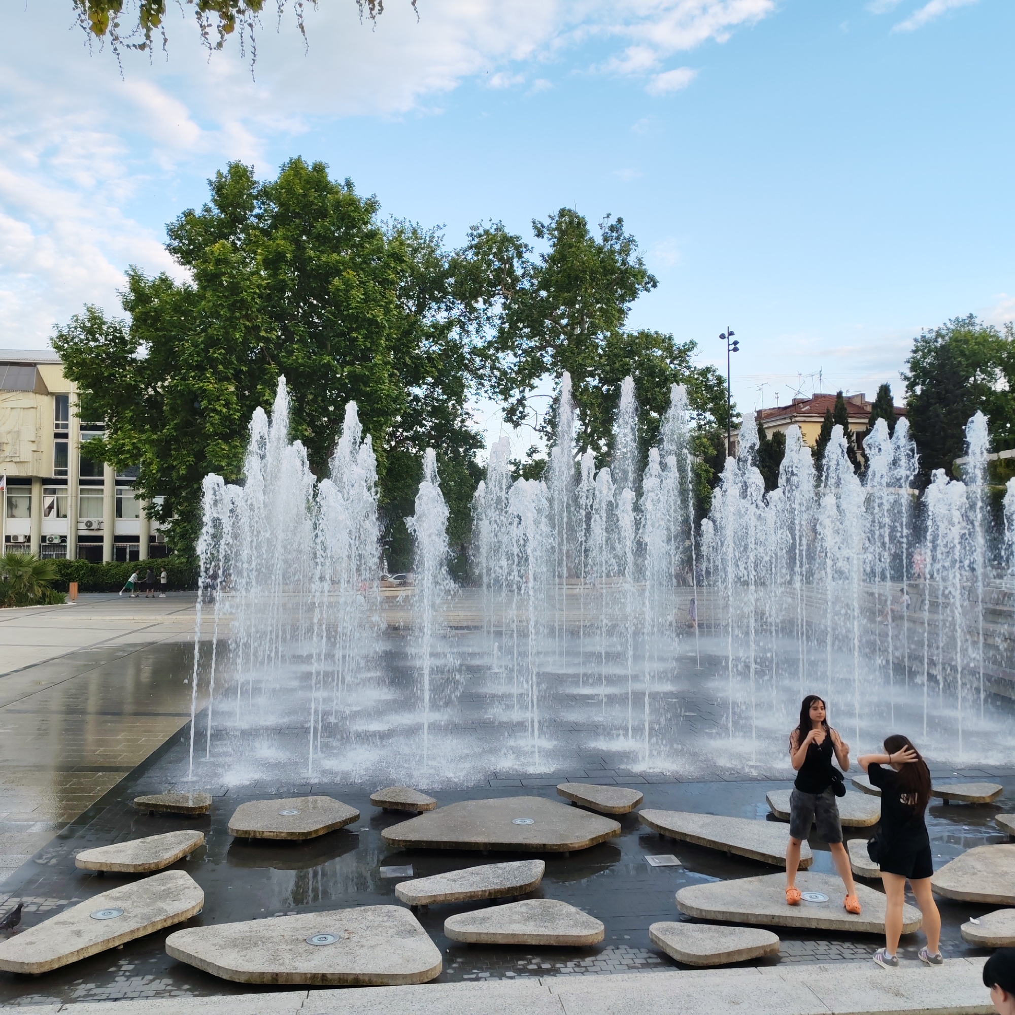 This image depicts an outdoor scene featuring a large fountain with multiple jets of water spraying upwards in a public square. In the foreground, two young women are standing on flat, irregularly shaped stone platforms within the water feature. The woman on the left is wearing a black tank top and grey shorts with orange clogs, while the woman on the right is wearing a black t-shirt and black shorts with grey and white athletic shoes. Both appear to be interacting with each other, with the woman on the right touching her hair.
The fountain is situated in front of a modern building with a light-colored facade and multiple windows. Behind the building and the fountain are large, mature green trees. The sky above is blue with scattered white clouds. The ground of the square is paved with light-colored tiles, and the area around the stone platforms is wet from the fountain's spray, reflecting the water and sky. The presence of architectural elements suggests an urban environment. The image seems to be taken during daylight hours.