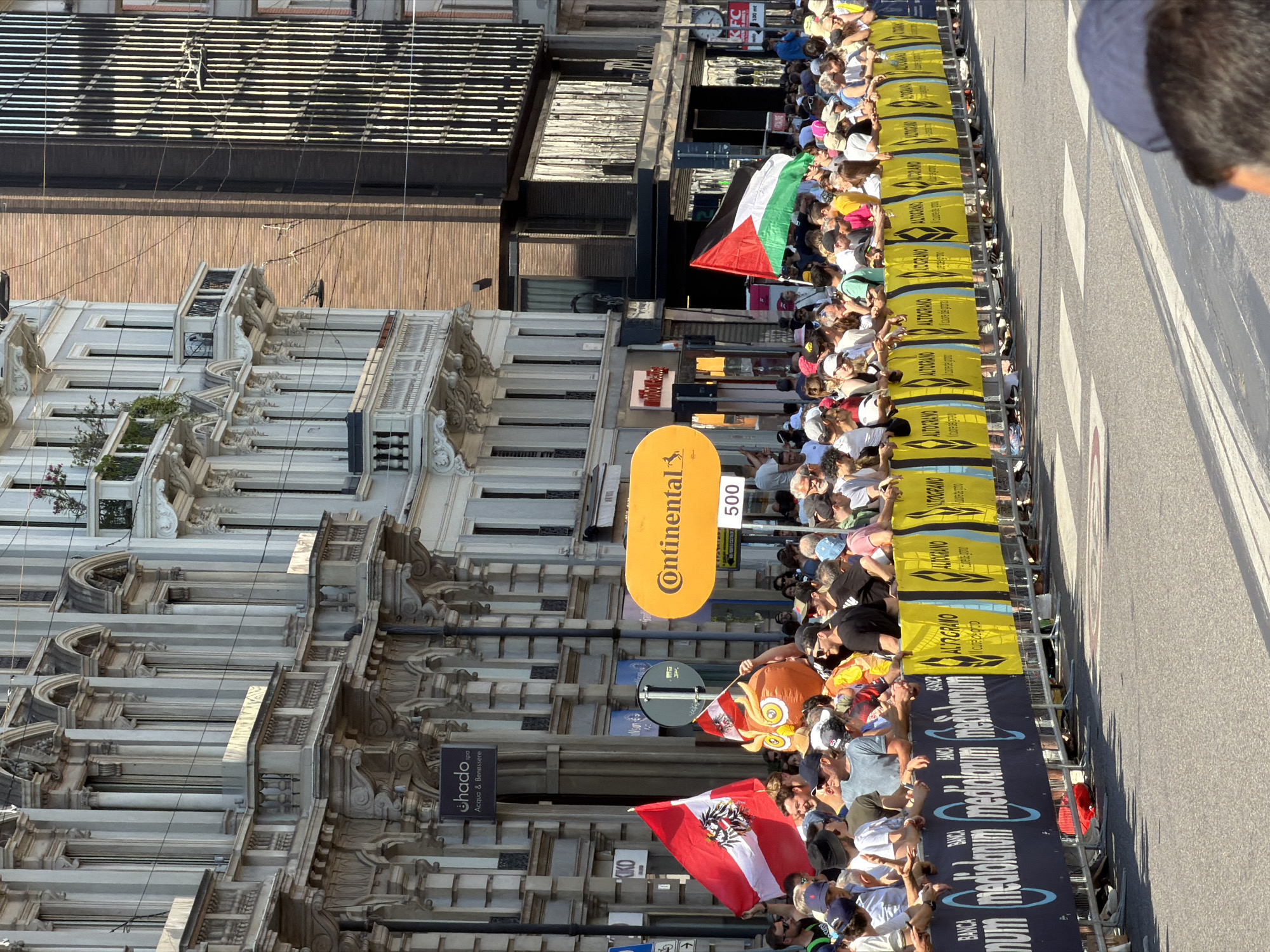 The image shows a street scene in Milan, Italy. A large crowd is lining the street behind barricades to watch an event, likely a bicycle race, as evidenced by the "Continental 500" sign and flags being waved. The barricades are decorated with advertising for "ALTOGRANO" and "BANCA MEDIOLANUM." Flags visible include those of Austria and Palestine. On the left side of the frame, there are ornate, older buildings with detailed architectural features. Street signs and shop signs, including "hado Acqua & Benessere" and "KIKO," are also visible on the buildings. In the background, a clock and signage reading "KFC" can be seen above the crowd. The street itself is marked with zebra striping, indicating a crosswalk. The overall scene is bright and sunny.
