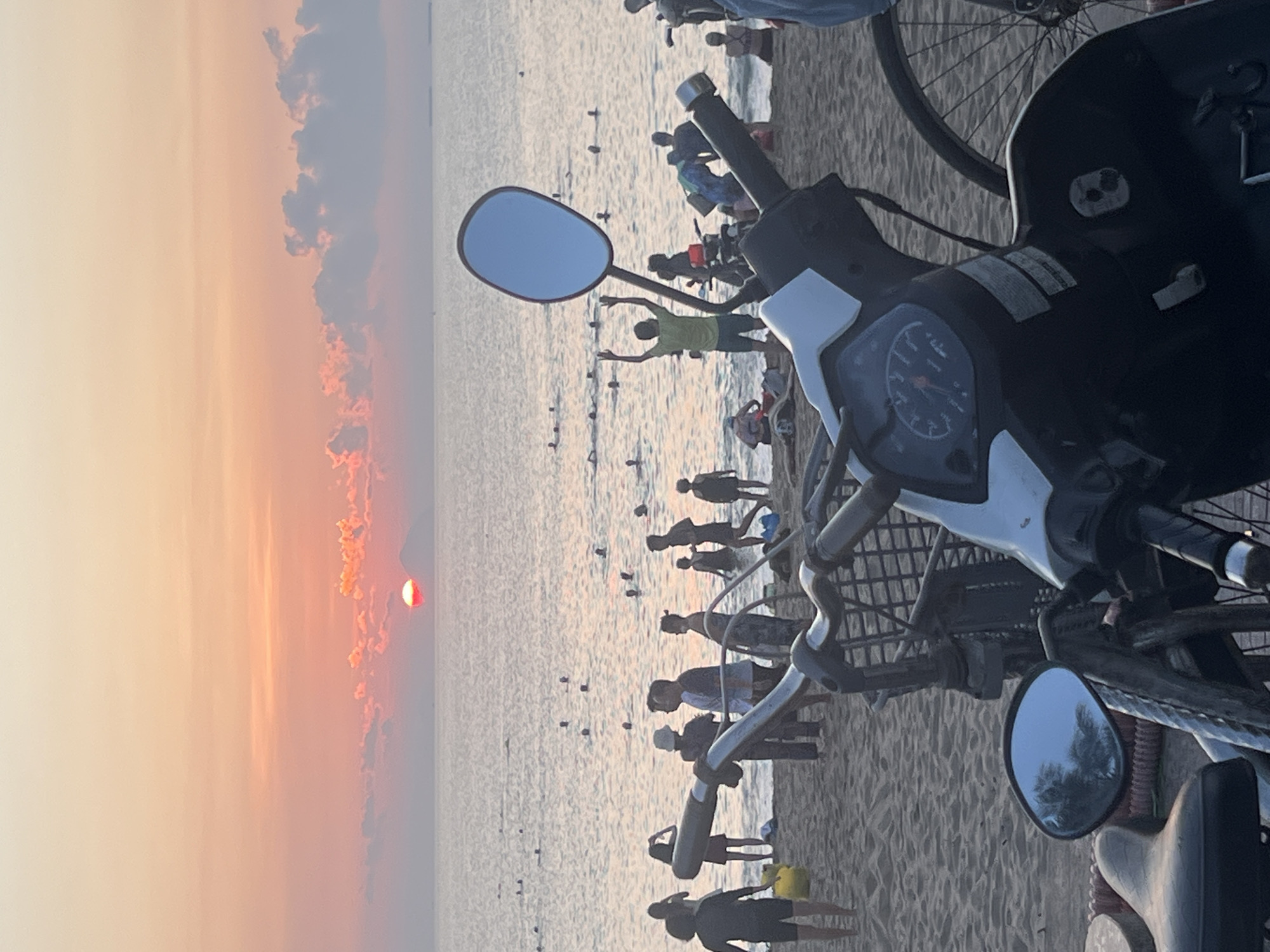 The image depicts a beach scene, likely in Nha Trang, Vietnam, at sunset. The sun is setting on the left side of the frame, casting an orange and pink hue on the sky and water. People are on the beach, some in the water, some walking along the shore. There is a motorcycle parked on the beach, partially obscuring the view of the sunset and the people.