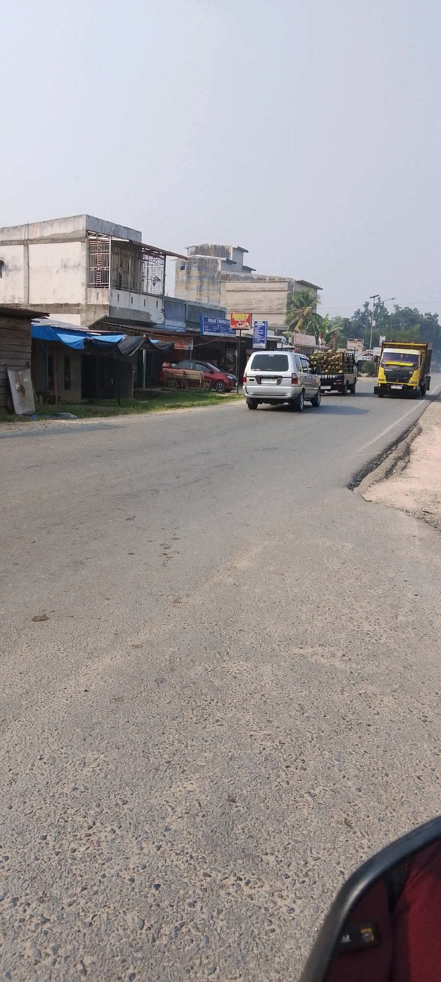 The image shows a street scene in what appears to be a commercial area. A paved road runs diagonally from the lower left to the upper right of the frame. On the left side of the road, there are several single-story buildings with corrugated metal roofs and awnings. One of these buildings has a blue tarp covering part of its frontage. Further back, there are multi-story buildings with concrete facades and balconies. A red car is parked in front of one of the buildings.
On the road, there are several vehicles. A silver SUV is in the foreground, moving away from the camera. Behind it, a truck carrying what appears to be coconuts is also moving away. To the right of the coconut truck, a yellow dump truck is also traveling in the same direction. The sky is overcast with a light haze. The general impression is of a somewhat undeveloped commercial strip on a moderately busy road. The location is identified as Seremban Jaya, Indonesia.