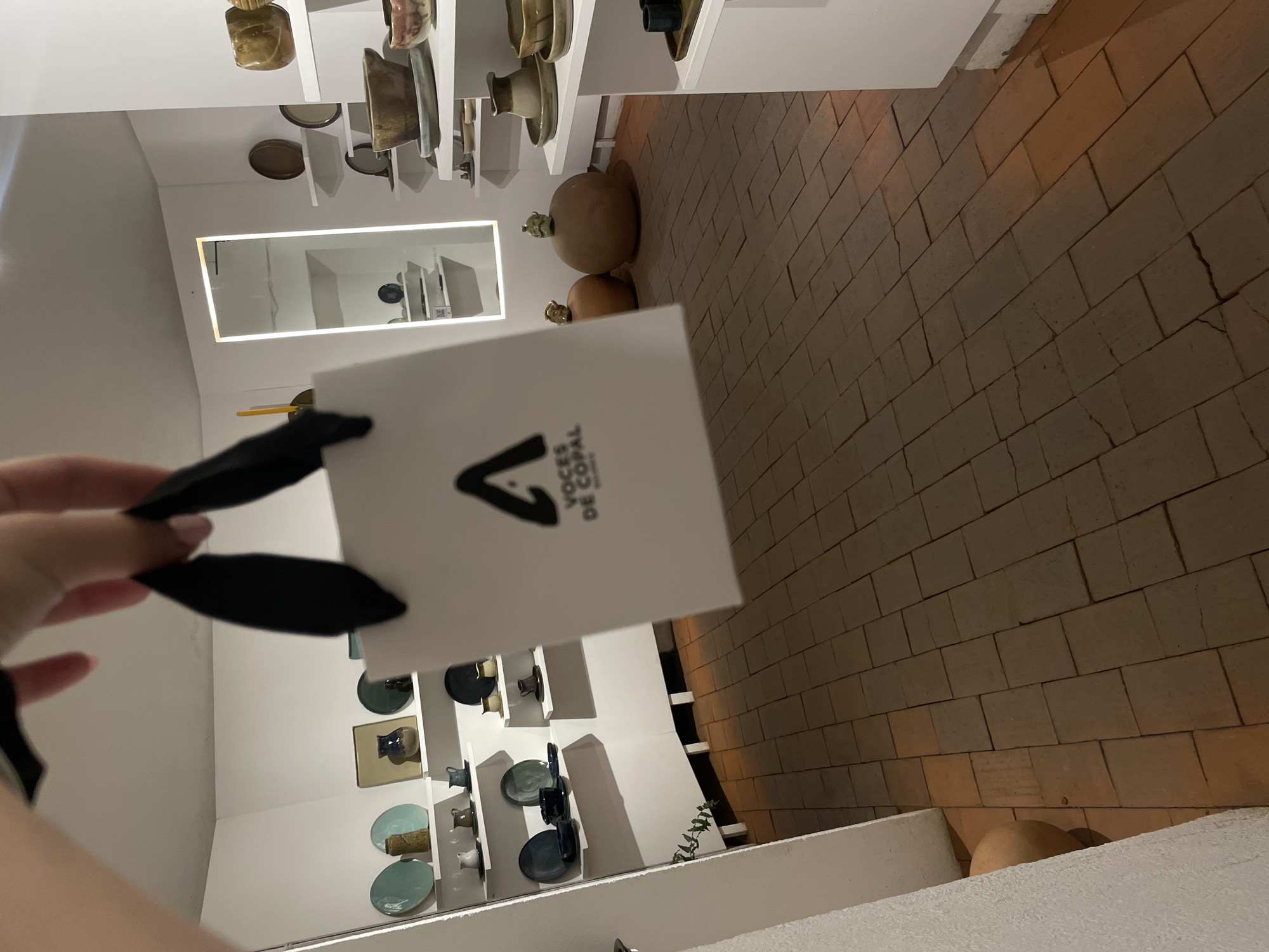A person holds a white shopping bag with the "Voces de Copal" logo, against the backdrop of what appears to be an art gallery or shop interior in Oaxaca City, Mexico. The bag's black ribbon handles are visible, and the logo is a stylized upward-pointing arrow above the name.
The background includes a display of pottery and artisanal objects on white shelves against white walls. There's a mirrored section reflecting the shelves. The gallery space leads to a brick-tiled floor. Large, round ceramic objects sit on the floor, likely part of the gallery's collection. The scene suggests the person has made a purchase from Voces de Copal, a business associated with copal wood carvings, and is inside or just outside their shop in Oaxaca City.