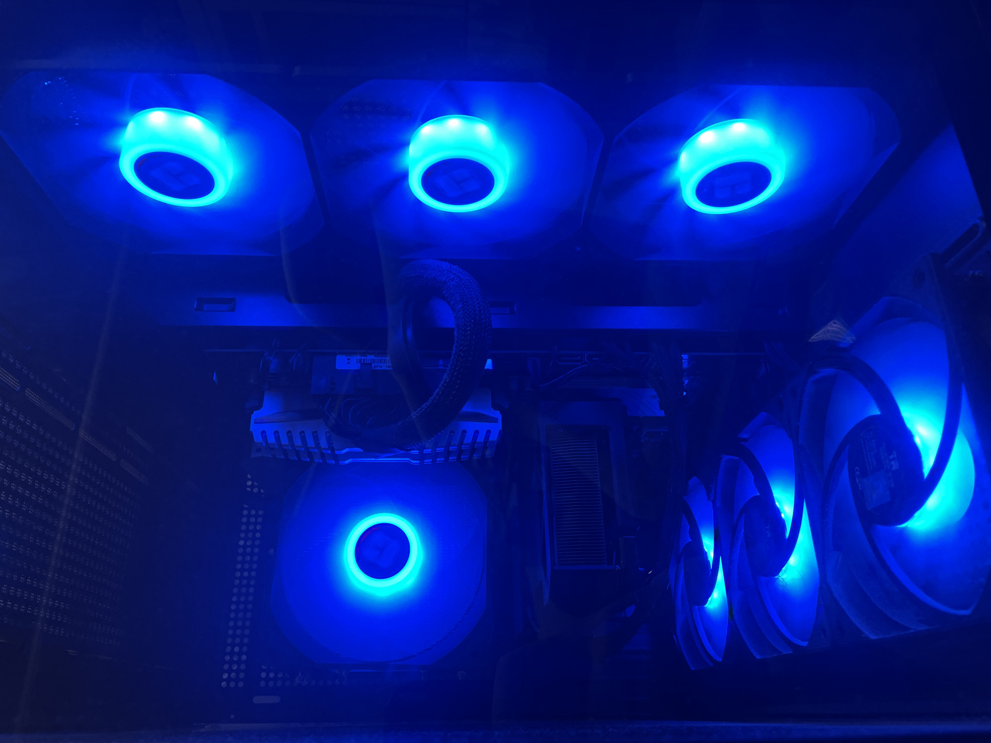 The image shows the interior of a computer case, illuminated by blue LED lights from multiple fans. There are several fans visible: one large fan at the top left, another large fan at the top right, and three smaller fans stacked vertically on the right side of the case. A heat sink is visible in the lower left. The overall environment is dark, with the blue lights creating a dramatic effect. The image is a close-up view of the computer's internal components. There is no indication of location in a city or country.