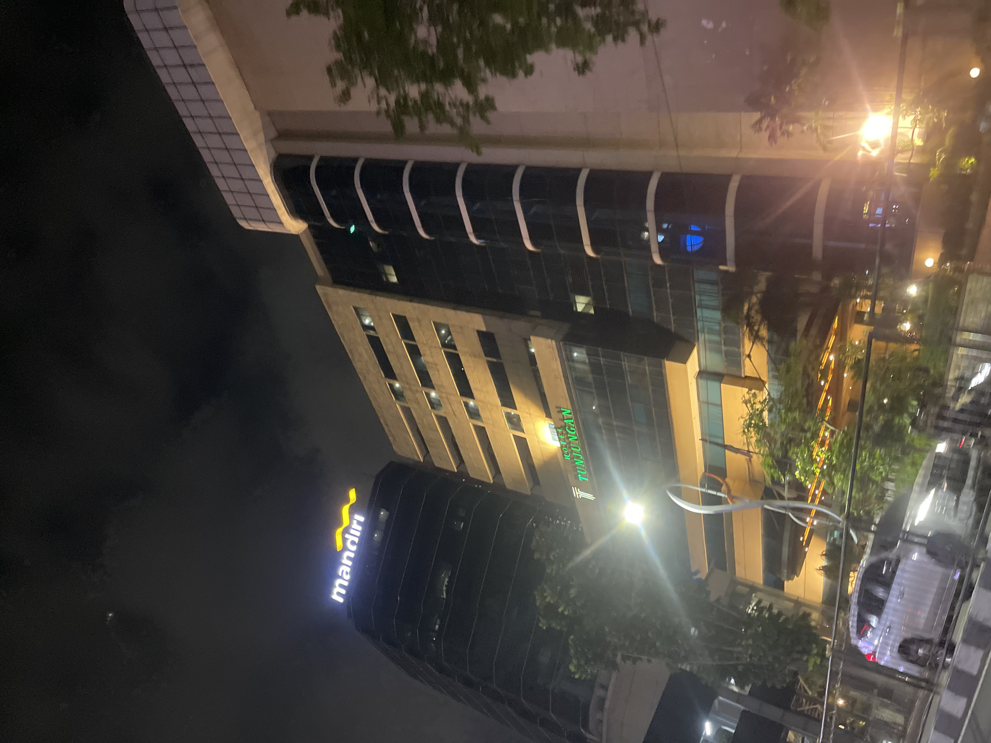 This is a nighttime, low-angle exterior shot of two adjacent buildings in Surabaya, Indonesia. The building on the left is a dark, modern skyscraper with a prominent illuminated "mandiri" logo at its peak. The building to its right appears older, with a lighter facade and rows of windows. Below these buildings, a section of a hotel is visible, displaying an illuminated sign with a stylized "T" logo and the text "TUNJUNGAN". In the foreground, a silver car is parked on the street, partially obscured by a metal railing and trees. Streetlights and building lights create a dynamic interplay of illumination and shadow, with lens flares from bright lights affecting parts of the image. The sky is dark with some wispy cloud formations.