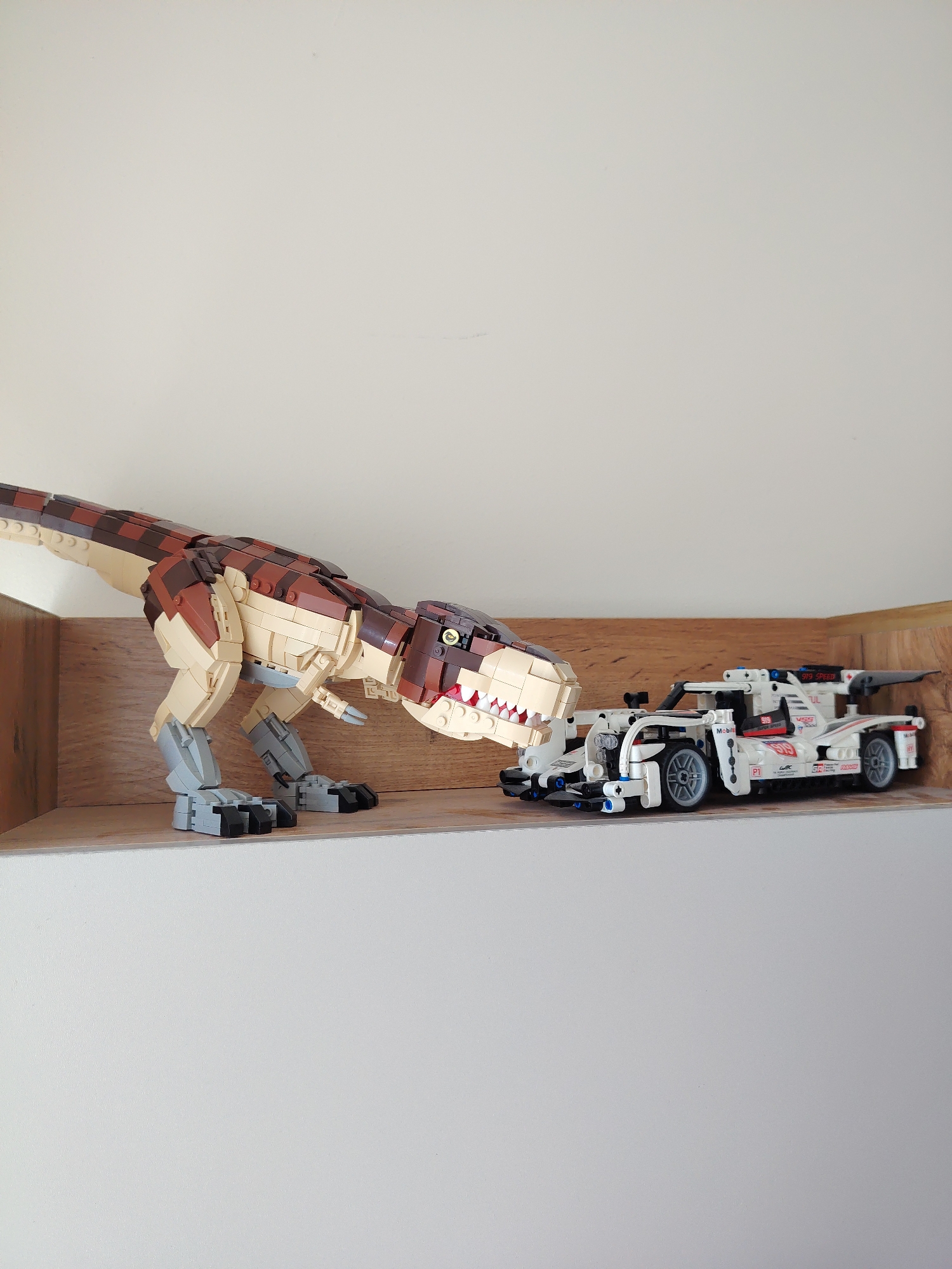 A Lego Tyrannosaurus Rex and a Lego race car are positioned on a wooden shelf against a white wall. The T-Rex, constructed with tan and brown bricks, is positioned to the left, with its head facing towards the race car. The race car, a white model with red and blue accents, is to the right, and appears to be a Porsche 919 Hybrid. The scene suggests a playful interaction between the two toys. The image does not contain any information about the city of Angren, Uzbekistan.