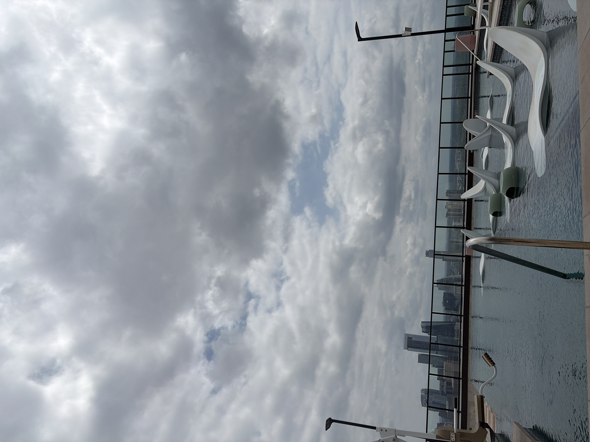 This image presents a high-angle, panoramic view from what appears to be a rooftop or elevated terrace, showcasing a city skyline under a dramatic, cloudy sky. The scene is likely situated in Austin, United States of America, as per the location context.
The dominant feature in the foreground is a large, rectangular body of water, resembling a swimming pool or reflective pond. Several strikingly modern, white, abstract sculptural pieces, possibly loungers or decorative elements, are placed within or along the edge of this water feature. Their organic, flowing shapes contrast with the rigid lines of the urban background. A transparent glass and metal barrier runs along the far edge of the water, separating it from the distant cityscape.
Beyond this barrier, a dense urban skyline emerges, featuring numerous tall, contemporary buildings that stretch across the horizon. The architectural styles suggest a modern metropolis.
The sky occupies a significant portion of the image, filled with a dynamic mix of dark gray and bright white clouds, indicating an overcast day. Patches of lighter, almost blue sky are visible through breaks in the cloud cover, suggesting diffused daylight rather than direct sunlight. Two black, L-shaped poles, possibly part of lighting fixtures or structural elements, extend into the sky from the periphery of the frame. No discernible human activity or visible text is present, lending a serene and contemplative atmosphere to the urban landscape.