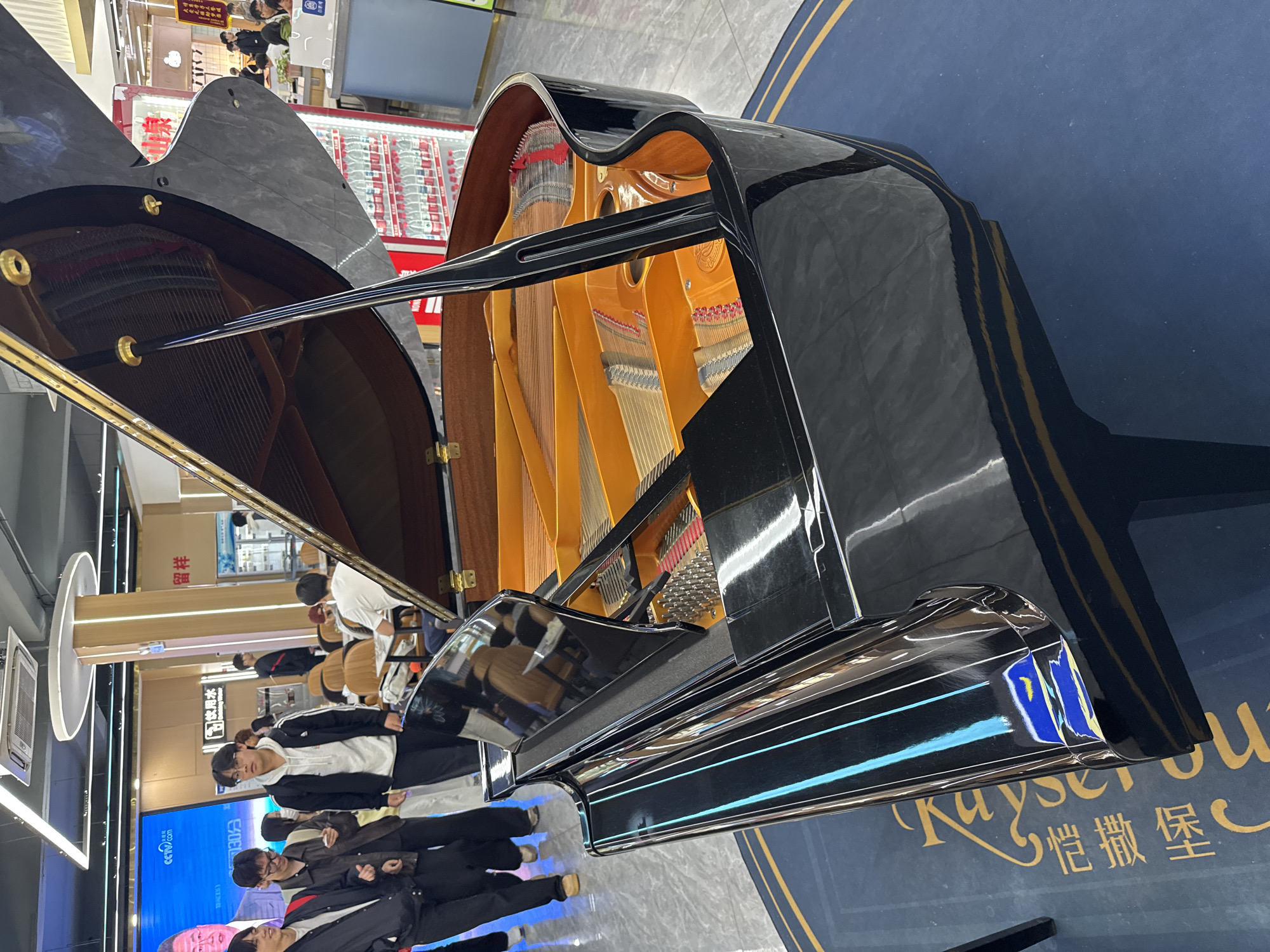 A black, highly polished grand piano is prominently displayed in the foreground of this image, set indoors within what appears to be a modern shopping mall or large retail space in Zhongtun, China. The piano's lid is propped open, offering a view of its internal components, including the warm, yellowish wooden soundboard, numerous strings, and hammers. Golden hardware, such as hinges and pins, accentuates the instrument's design. Its glossy surface reflects the bright overhead artificial lighting and elements of the surrounding environment.
The piano is positioned on a large, circular blue mat adorned with decorative gold lines. Visible text on the mat clearly states "Kayser" in a cursive English font, followed by the Chinese characters "愷撒堡," identifying the brand as Kayserburg.
Multiple individuals, predominantly young adults or teenagers in casual attire, are seen standing around the piano and in the background. Some appear to be observing the instrument with interest, while others are simply passing through the area. No one is actively playing the piano.
The background reveals a typical commercial setting with several brightly lit shopfronts and stalls. Reflections on the piano's surface show modern strip lights and circular light fixtures on the ceiling. A white wall-mounted air conditioning unit is visible on the left. Additionally, a screen in the background displays "CCTV.com," indicating content from the Chinese national broadcaster. The scene conveys a casual, public atmosphere during what appears to be daytime or evening, given the reliance on artificial lighting.