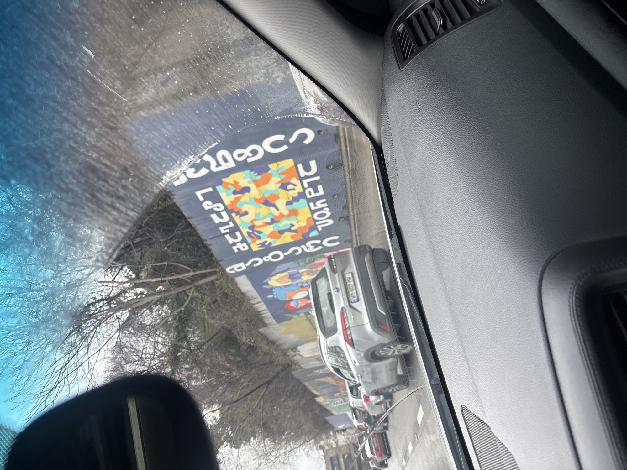 The image captures a street scene from the interior of a vehicle in Tbilisi, Georgia, looking through a rain-splattered windshield. The dark gray dashboard and a portion of the rearview mirror are visible in the foreground. A line of vehicles is present on a multi-lane asphalt road, suggesting traffic congestion. Directly ahead is a silver Toyota Corolla Cross SUV with the Georgian license plate "WM-007-WW." To the right of the road, a long retaining wall, recognized as the Georgian Alphabet Wall, features vibrant murals with abstract colorful patterns and Georgian script, including "ქართული ანბანი" (Georgian Alphabet). Above the wall, bare deciduous trees and vegetation cover a sloping embankment under an overcast sky. This wall is situated along the Mtkvari River embankment, consistent with sections of Akaki Tsereteli Avenue or David Agmashenebeli Avenue. No people are visible. The vehicles appear to be stationary or moving slowly.