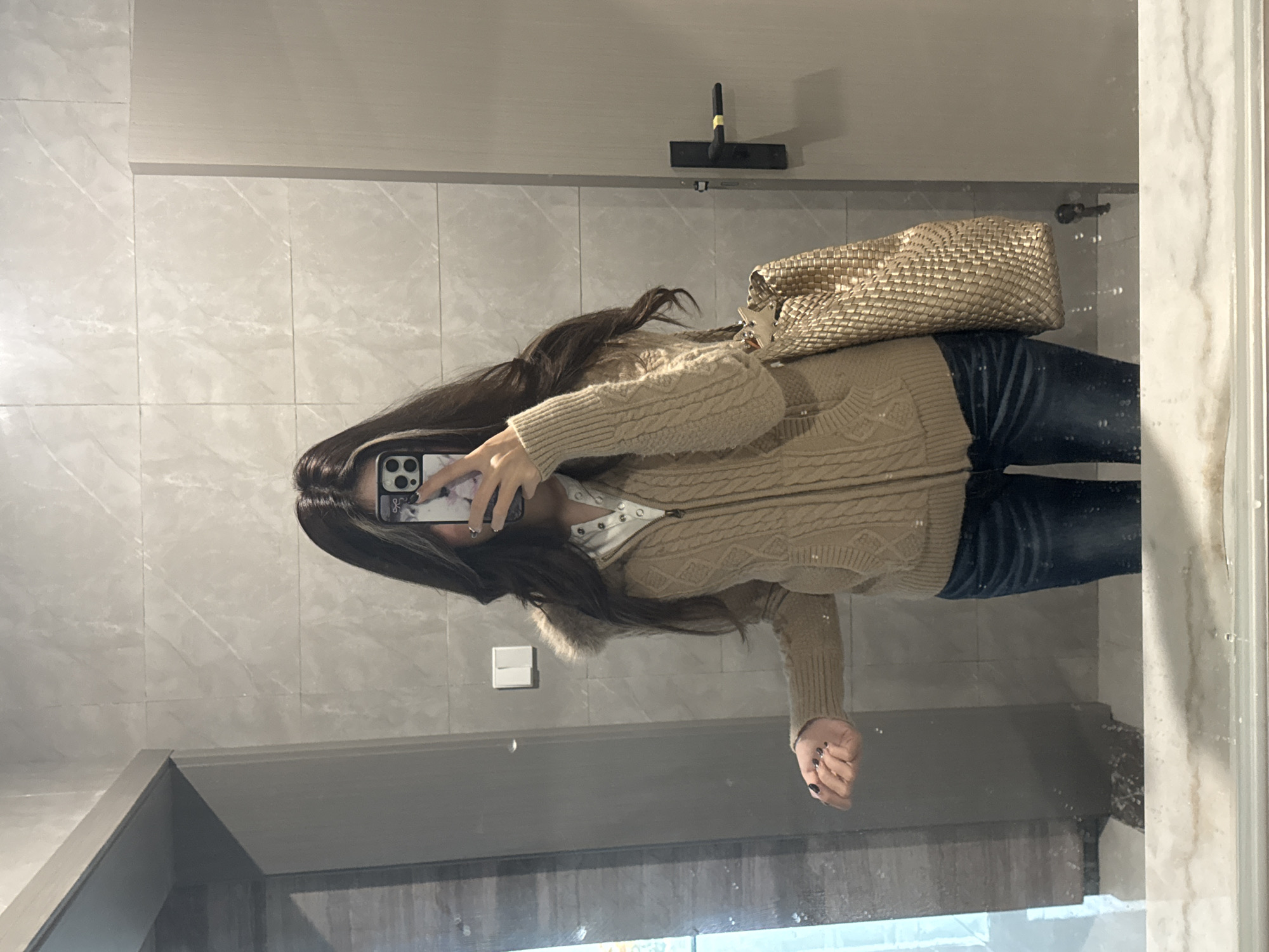 The image displays a mirror selfie of an individual taken indoors. The location is specified as Nan'an District, China. A single person is visible, primarily from the chest to the mid-thighs, with their face obscured by a smartphone held by their right hand. The person has long, dark hair with a lighter-colored streak on the right side (viewer's right). They are wearing a beige cable-knit hooded cardigan with visible fur trim on the hood, layered over a white collared shirt with a button placket. Dark blue jeans cover the lower body. The left hand is extended slightly, showing dark nail polish. A metallic, gold-colored, woven-texture shoulder bag hangs on the person's left side. The interaction involves the individual taking a photograph of their reflection using a smartphone with a patterned case. The environment consists of an indoor room with light gray rectangular wall tiles, some exhibiting subtle marbling. Reflected in the mirror are a white rectangular light switch with two buttons, a light-colored door with a black handle featuring a yellow detail, and a section of a light-colored counter or surface below the mirror, which shows water marks or splashes.