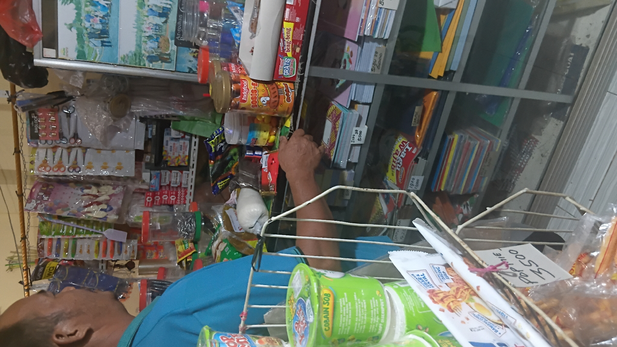 The image shows a man in a blue shirt standing in front of a store display. The display contains various items such as snacks, stationery, and other small goods. The store appears to be a small, local shop in Prabumulih, Indonesia, likely a "warung" or convenience store. The man is facing away from the camera, his left arm is extended towards the shelves. The shelves are made of metal and glass, and are filled with neatly organized products. The overall lighting suggests an indoor setting, possibly with artificial light. The image captures a moment in a typical Indonesian small business environment.