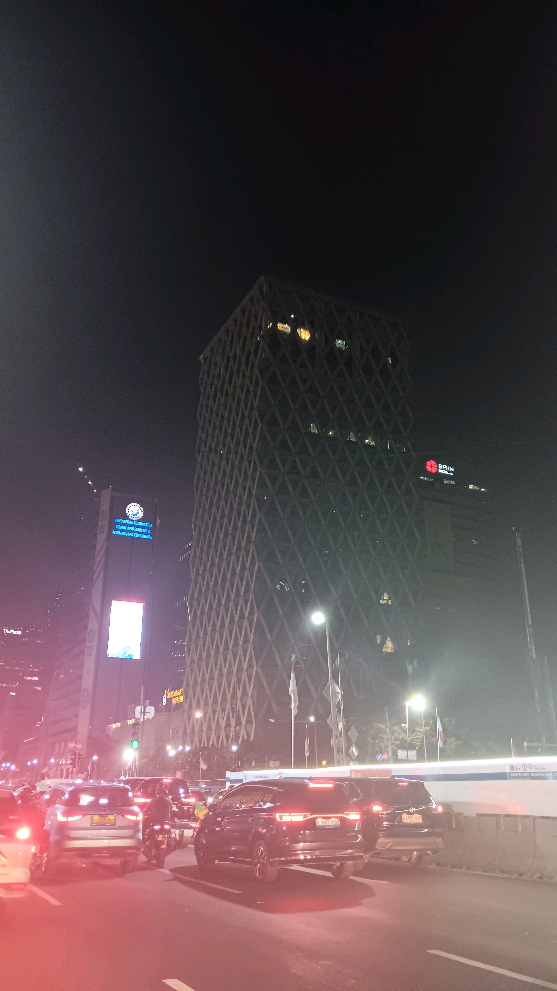 The image depicts a nighttime street scene in Central Jakarta, Indonesia, characterized by heavy traffic. A prominent, angular skyscraper with a patterned facade dominates the background, illuminated by internal lights. Adjacent to it, another taller building displays a blue illuminated sign that reads "KEMENTERIAN KOORDINATOR BIDANG PEMBANGUNAN MANUSIA DAN KEBUDAYAAN" and a circular emblem. To the right, a smaller building shows the red logo of "BRIN". The foreground is filled with a gridlock of vehicles, including cars and motorcycles, their headlights and taillights creating streaks of light. The overall atmosphere is one of urban congestion under artificial illumination, with a slight haze or fog present in the air.