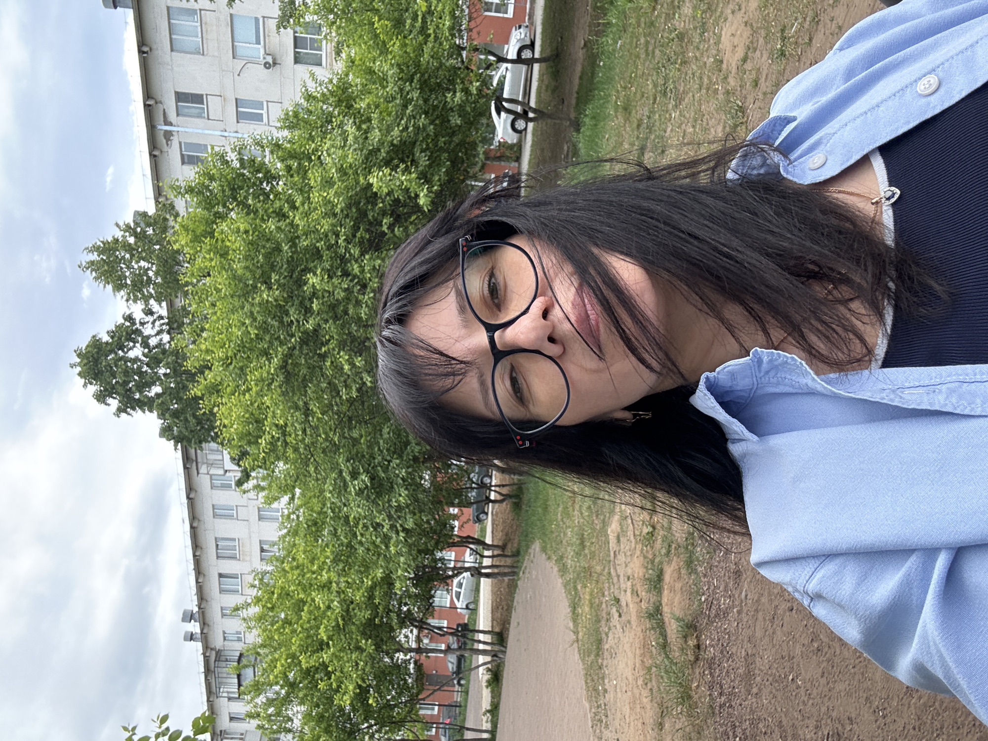 A woman wearing glasses and a light blue shirt is outdoors. A building with multiple windows is visible in the background behind trees. A sidewalk is present in the foreground and a car can be seen parked in the distance.