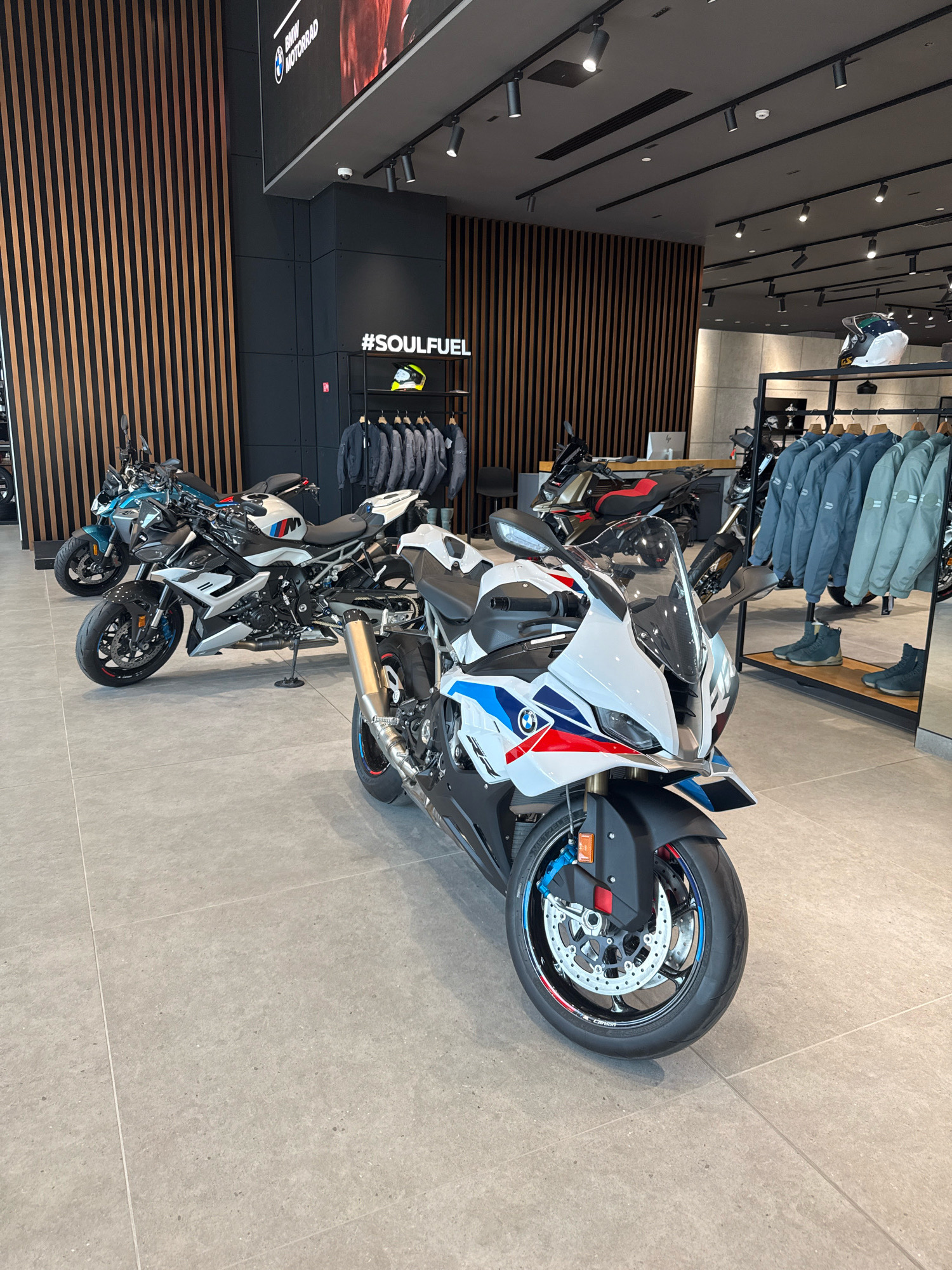 The image depicts the interior of a BMW Motorrad dealership in Dubai, United Arab Emirates. Several motorcycles are on display, with a prominent white and blue BMW S1000RR sports bike in the foreground. To the left, two other BMW motorcycles, one a teal and white model and another a black and red model, are visible. In the background, a rack of BMW motorcycle jackets and helmets is displayed, along with a sign that reads "#SOULFUEL". The dealership has a modern aesthetic with wooden slatted walls and a grey tiled floor. The lighting appears to be artificial, coming from track lighting on the ceiling. There are no people visible in the image.