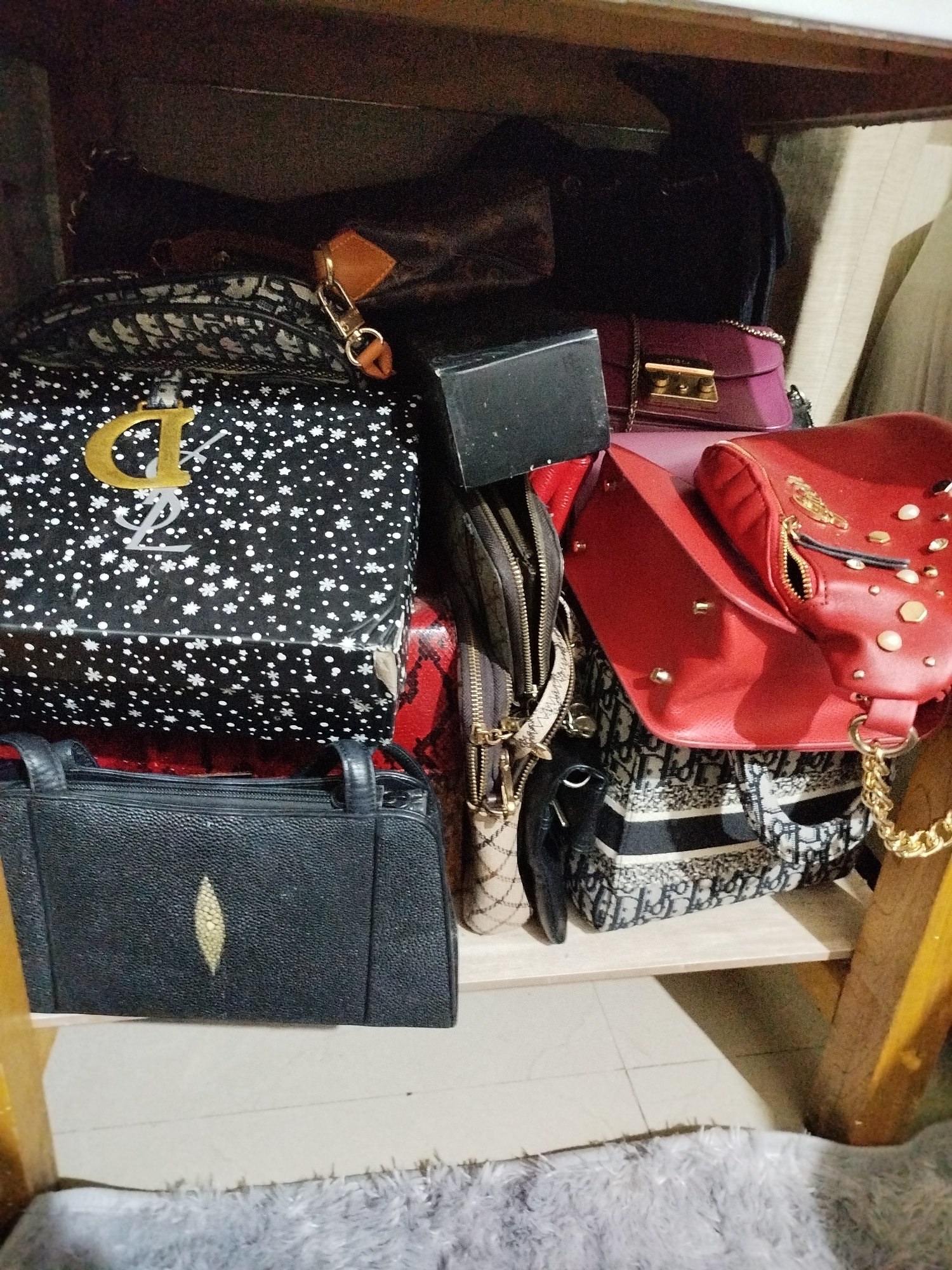 A collection of luxury handbags is displayed on a wooden shelf. From top to bottom, left to right, the handbags include a dark-colored bag with a tan leather tag, a black and white patterned bag with a large gold "YSL" logo, a red handbag with gold stud embellishments, a black textured handbag with a light-colored oval detail, and a large patterned tote bag with a repeated logo design. The environment appears to be an indoor setting, possibly a closet or retail space, with a glimpse of a fluffy grey rug in the foreground. The image does not contain any identifiable location markers for Bandungrejo, Indonesia.