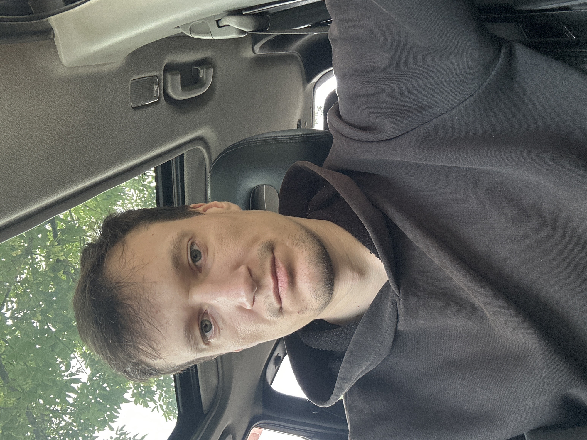 A man is taking a selfie inside a car. The car's sunroof is open, revealing a glimpse of green trees and a cloudy sky above. The man is wearing a dark hoodie and has short brown hair. He is looking directly at the camera. The interior of the car is visible, including the headrest of a passenger seat and part of the car's ceiling. The image was taken outdoors on a bright day. There is no indication of the specific location in Makiv, Ukraine, beyond the general setting of a car.