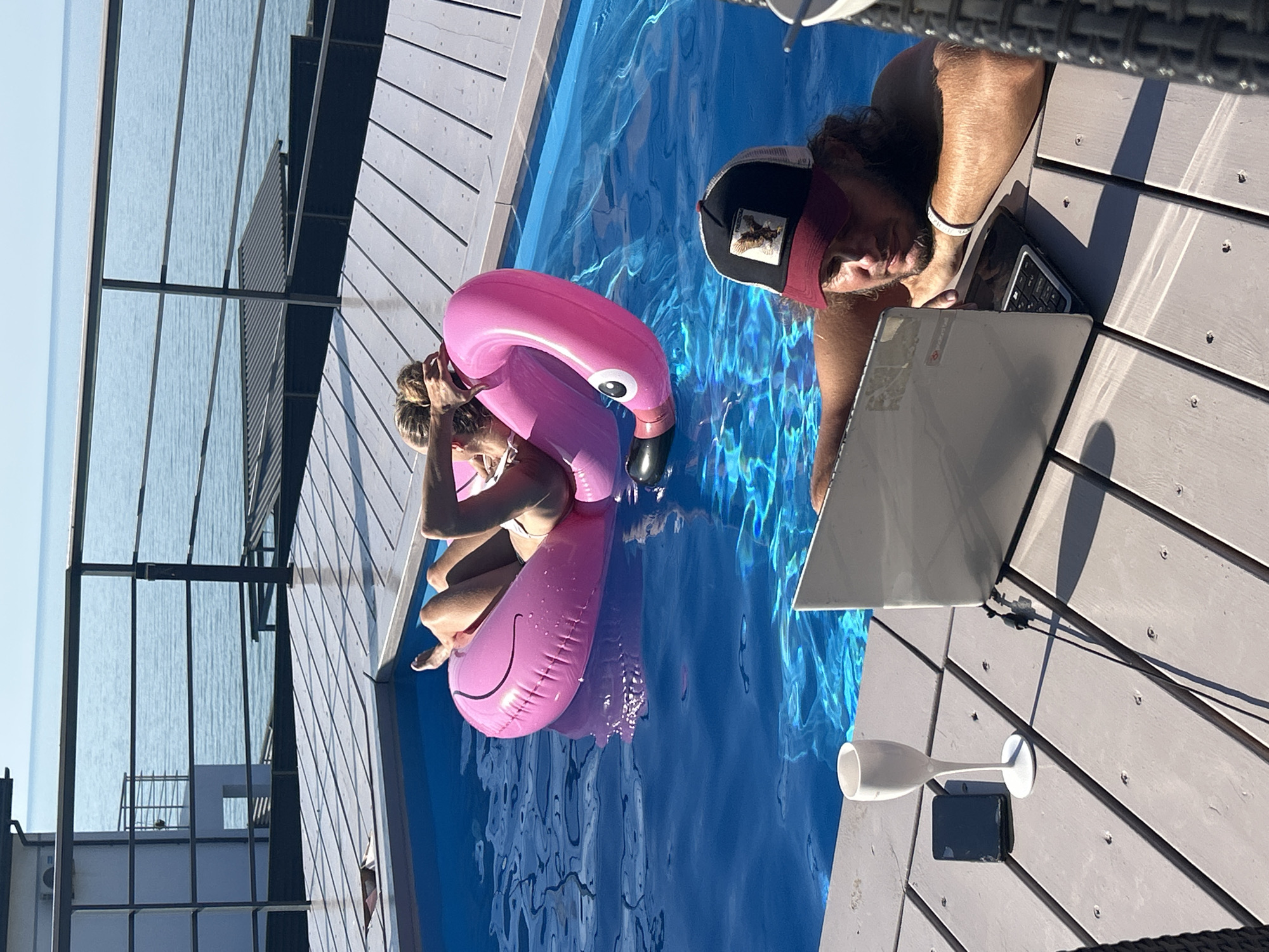 A person with dark curly hair, wearing a baseball cap and a white bracelet, is sitting by a swimming pool and looking at a laptop. The person is on a wooden deck, with a pool filled with blue water to their left. The person's right arm is resting on the laptop, and their left hand is near their face. A pink inflatable flamingo float is in the pool, near another person whose legs and torso are visible. This person is in the water, holding onto the flamingo, and appears to be sunbathing or relaxing. The scene is outdoors, suggesting a warm, sunny day, likely daytime given the bright lighting and clear blue sky reflected in the water. The background shows the calm, blue expanse of the sea or a large body of water. In the foreground on the deck, there is a white champagne flute and a dark object, possibly a phone or a small remote, placed next to the laptop. The wooden deck is made of grey planks. The overall atmosphere is one of leisure and relaxation, with a touch of work potentially being done despite the vacation-like setting. There is a visible logo on the laptop that reads "Lenovo". The cap worn by the person has a patch with an eagle or bird of prey.
