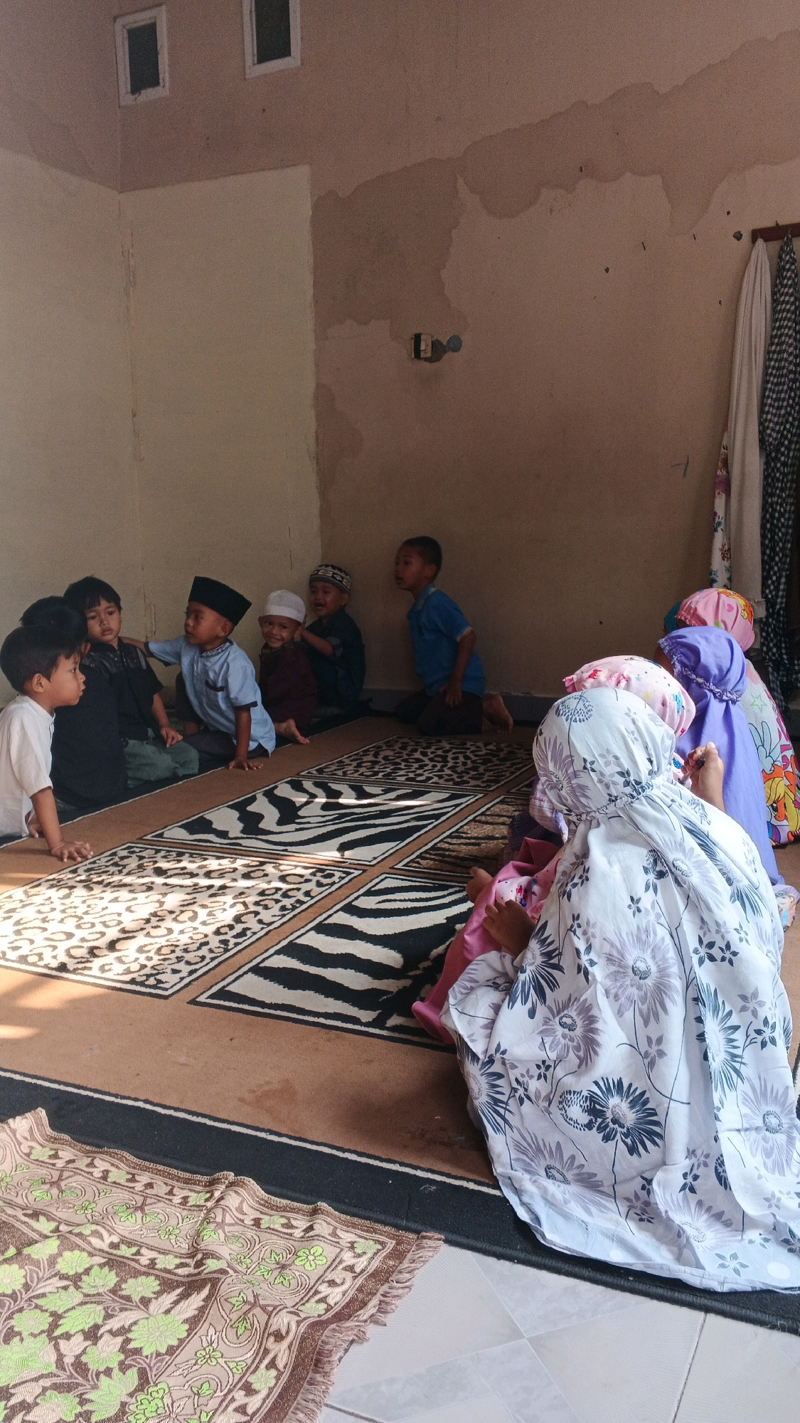 In a humble indoor setting, likely a home in Serang, Indonesia, a group of approximately ten young children are gathered on prayer mats. The children, appearing to be between the ages of four and eight, are dressed in modest attire, with several boys wearing skullcaps. Some are sitting cross-legged, while others are kneeling. The atmosphere suggests a religious gathering or educational session. The room itself is sparsely decorated, with neutral-colored walls and a tiled floor. Sunlight streams in, casting shadows on the patterned prayer mats, which are arranged in a rectangular formation. In the background, a door or opening can be seen, with some clothes hanging on a rack. The overall impression is one of a simple, community-focused activity in a modest residential environment within Serang.