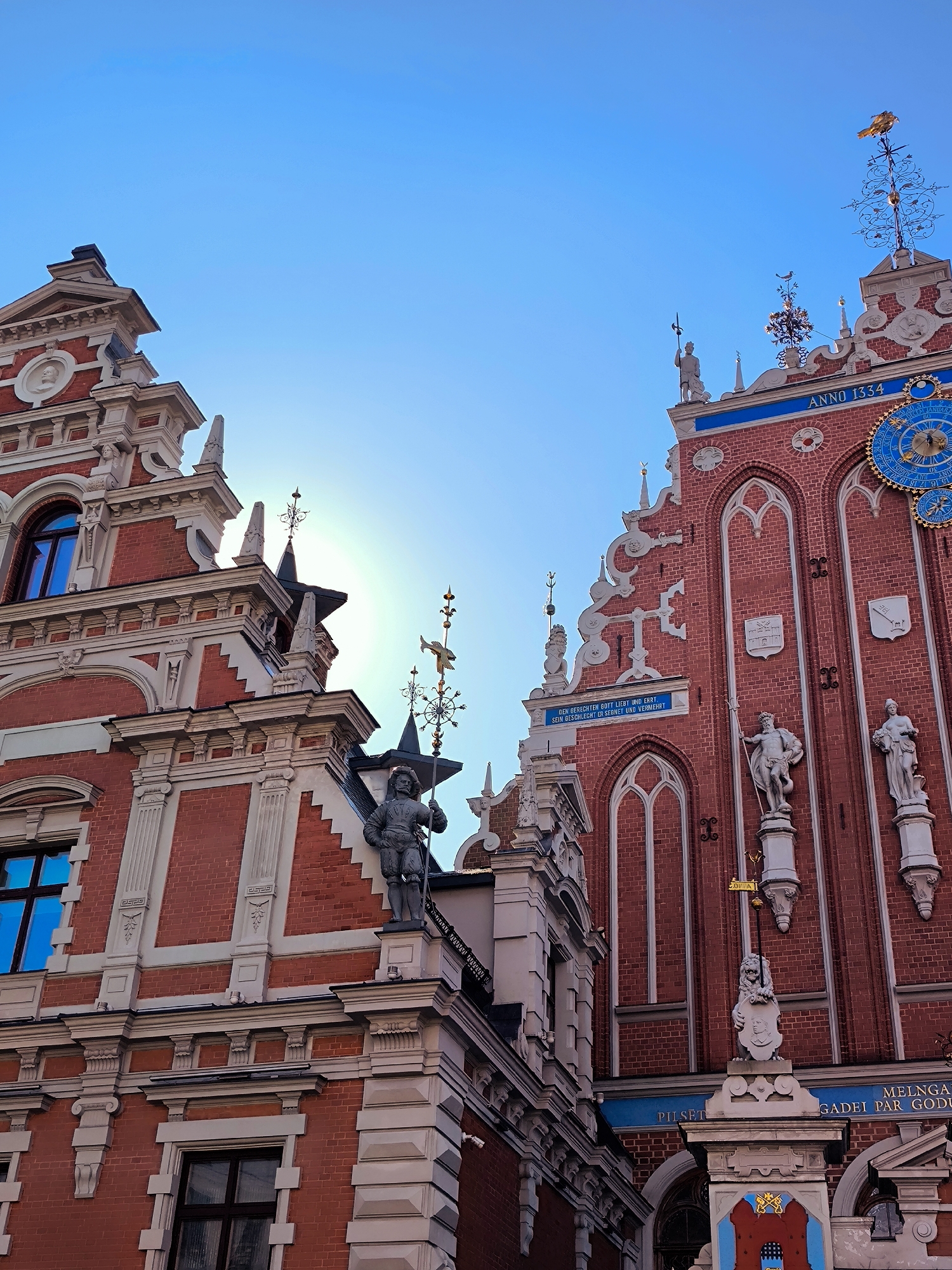 The image shows a close-up, low-angle view of the facades of two ornate red brick buildings in Riga, Latvia, against a bright blue sky with a visible sun flare. The building on the right is the House of the Blackheads, identified by the inscription "ANNO 1334" and a large blue astronomical clock on its facade. This building features Gothic-style arched windows, decorative sculptures of figures and mythical creatures, and a weather vane topped with a golden bird. The building on the left, likely a part of the same complex or adjacent, exhibits similar architectural details with red brick and white ornamentation, including decorative pilasters and a circular relief. The sun is positioned between the two buildings, creating a lens flare effect. The architecture is detailed, with intricate carvings and decorative elements characteristic of historic European buildings.