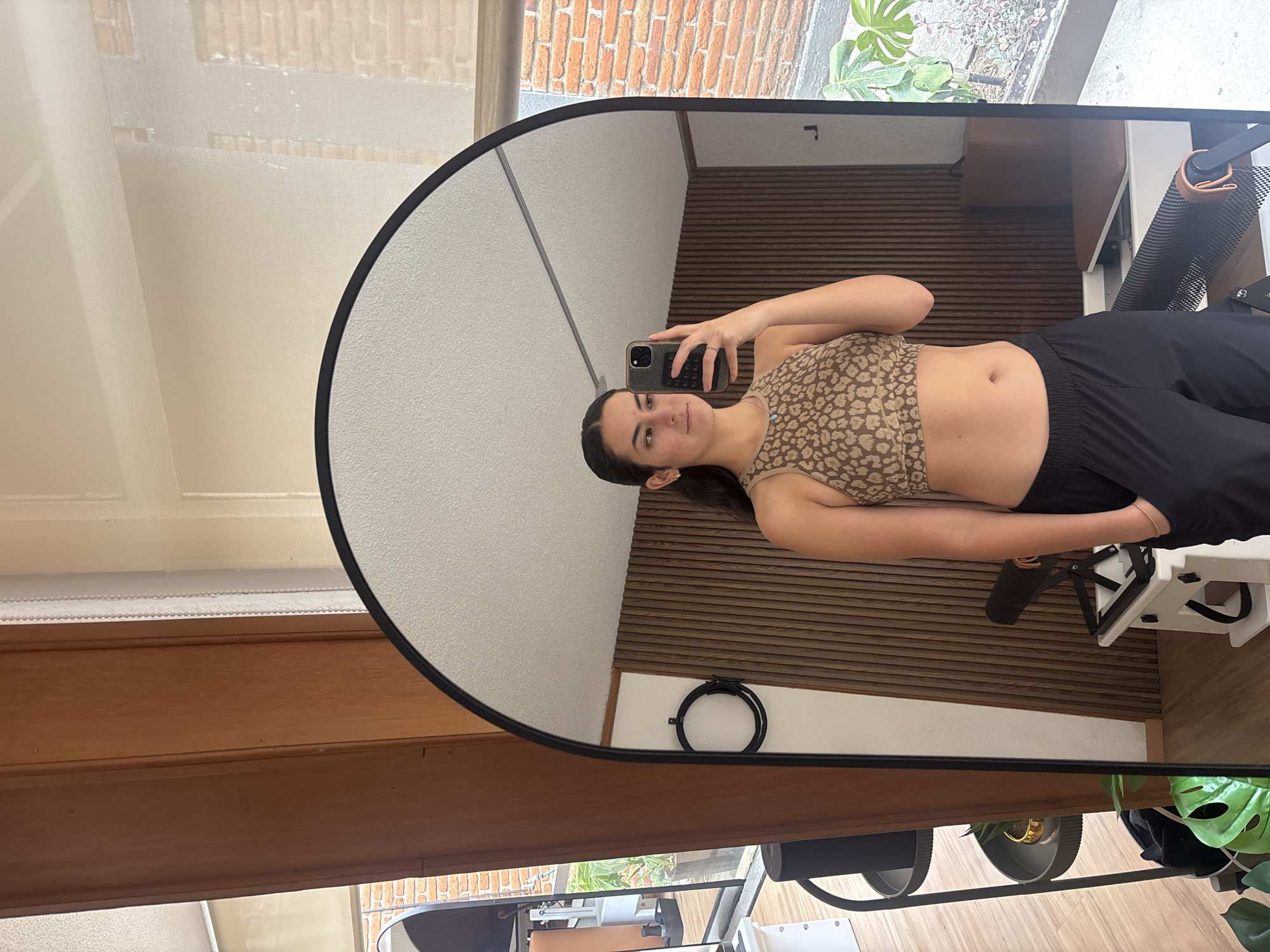 A woman is taking a selfie in a mirror. The woman is wearing a tan and brown leopard print crop top and black sweatpants. She has brown hair and is holding a black smartphone. The reflection in the mirror shows a room with white walls and wooden slat paneling, along with the workout equipment. The mirror reflects the room, revealing an exercise machine, possibly a pilates reformer. The location is in Puebla, Mexico.