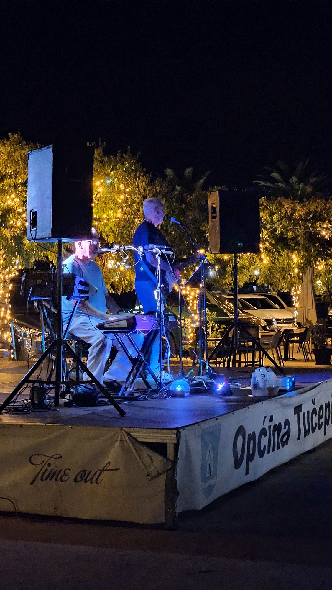 This image shows a nighttime outdoor performance in Općina Tučepi, Croatia. Two men are on a stage. The man on the left is seated and playing a keyboard. He is wearing glasses, a light blue short-sleeved shirt, and light-colored pants. The man on the right is standing and playing a guitar. He is wearing a dark blue t-shirt and dark pants. Both men are illuminated by stage lights. In the background, trees are decorated with numerous yellow string lights, creating a festive atmosphere. A white car is visible parked behind the stage. The front of the stage is draped with a banner that reads "Time out" in cursive on the left side and "Općina Tučepi" in block letters on the right side. A coat of arms is also visible on the banner. The ground in front of the stage appears to be paved.
