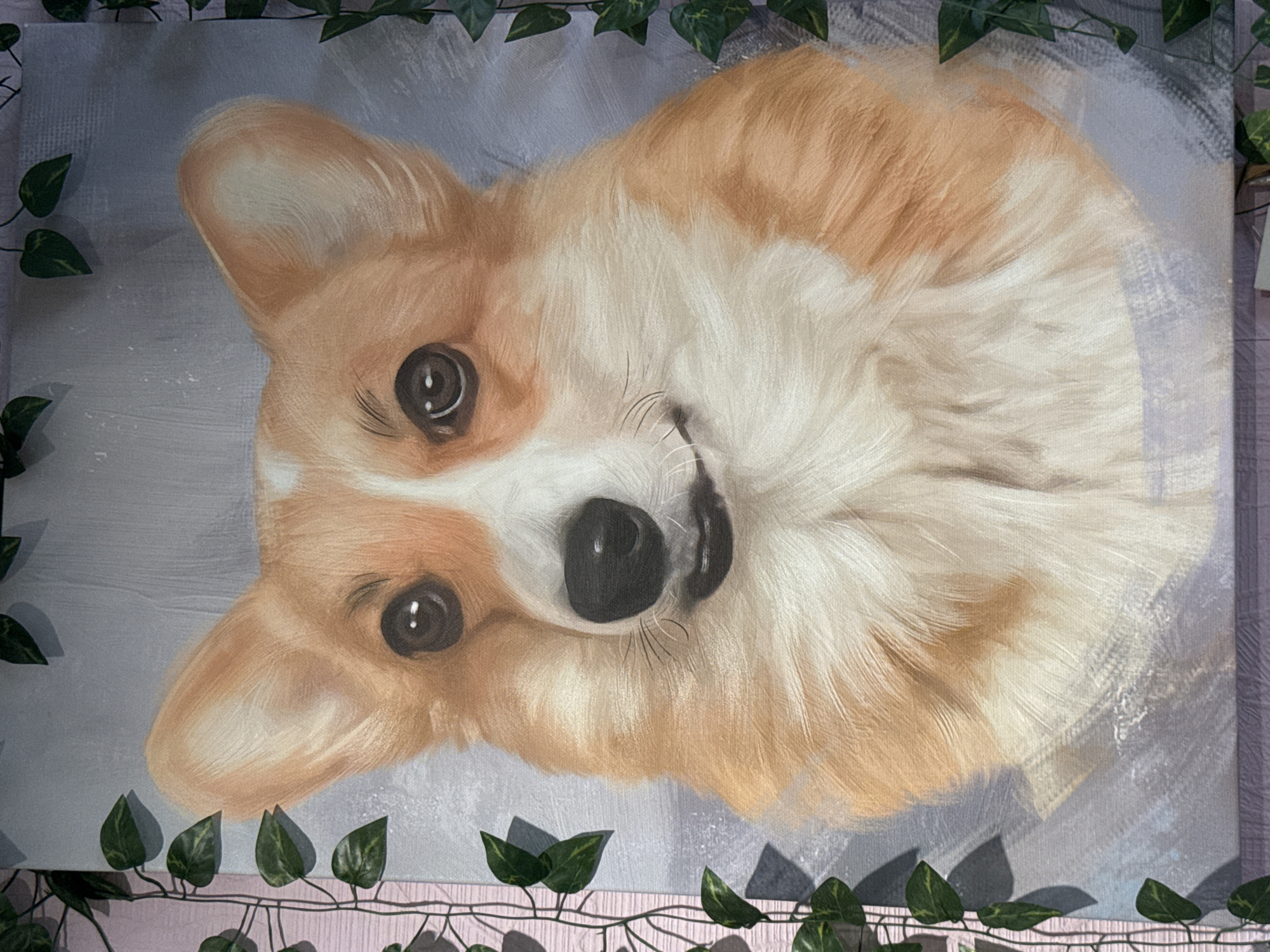 A close-up, vertical, eye-level painting of a corgi's face is shown. The corgi has large, expressive eyes, a wet black nose, and a fluffy cream and tan coat. The background is a soft, muted gray with hints of blue and purple. Green ivy vines are draped on either side of the painting, framing the corgi's face. The painting appears to be on canvas and is hanging against a pink, wooden-textured wall. A portion of a dark cord is visible at the bottom center of the image. The location is described as Unknown, Unknown.