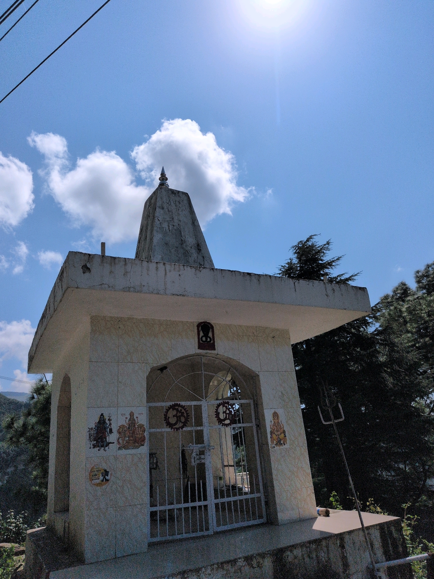 A small, white-tiled Hindu temple is situated on a slight incline, bathed in bright sunlight under a clear blue sky with scattered white clouds. The temple features a central arched entrance with a white metal gate, adorned with decorative motifs. Two images of deities, one depicting Shiva and Parvati and another of a single deity, are visible on the temple's facade, along with other decorative tiles. A tall, conical spire tops the structure. Surrounding the temple is lush green foliage and trees, with a hint of distant hills. The presence of power lines in the upper left corner suggests an inhabited area. The overall scene conveys a sense of peace and spirituality in a natural, mountainous setting, likely in Dharamkot, India.