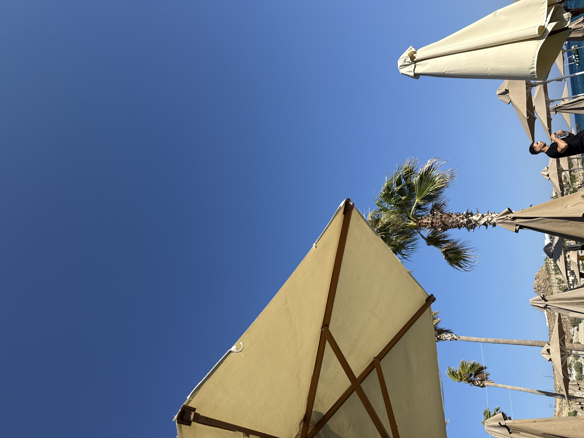 An outdoor, low-angle, full shot captures a sunny day on a beach in Plintri, Greece. The clear, vibrant blue sky dominates the upper portion of the image. In the foreground, a large, open, light-colored beach umbrella is angled upwards from the left, its wooden frame visible. Several other beach umbrellas, some open and some closed and covered, are scattered across the mid-ground. Palm trees, with their distinctive fronds, are interspersed among the umbrellas. A man in a black t-shirt is standing on the right side of the mid-ground, holding a water bottle and looking to his right. In the background, a rocky hill with sparse vegetation rises, and a few boats are visible on the bright blue water. The overall scene evokes a typical beach resort atmosphere.
