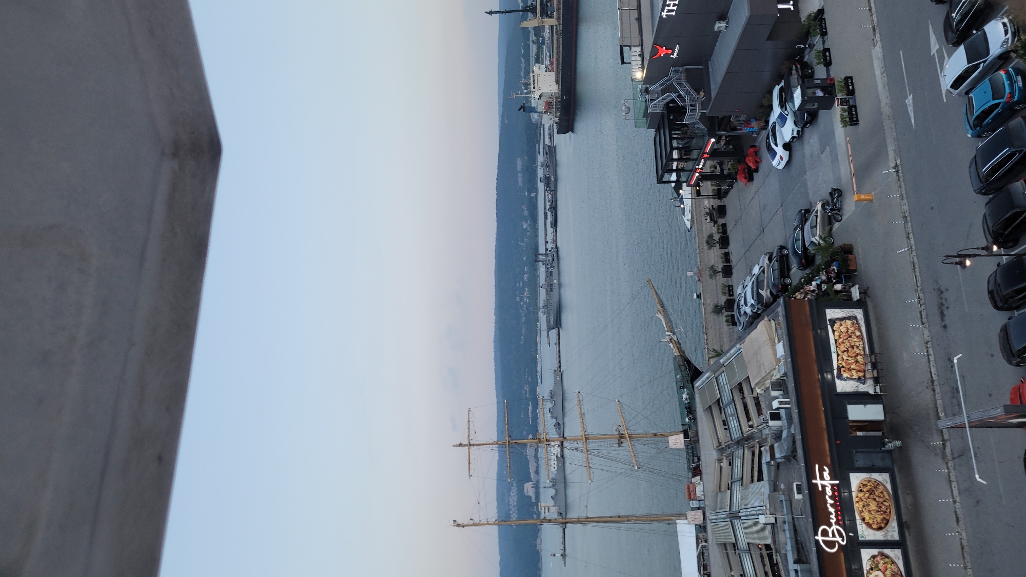 This media file, captured in Varna, Bulgaria, shows a waterfront scene at dusk. The composition is dominated by a large, grey, concrete structure in the foreground, taking up the left half of the frame. To the right of this structure, a calm body of water extends into the distance, lined by a tree-covered hillside.
On the waterfront, several objects and activities are visible. A tall-masted sailing ship is moored near the shore, its rigging prominent against the sky. Adjacent to the ship, a modern building with a dark facade features large windows and an outdoor seating area. The building has "Burrata" in large, stylized white lettering, suggesting it's a restaurant or pizzeria. Pictures of pizzas are displayed on the exterior. Several cars are parked along the street beside the building, and a few people are seated at tables outside.
Further along the waterfront, a large cargo ship is docked. The scene depicts a leisurely evening, with the soft twilight suggesting either early morning or late evening. The weather appears calm. The overall atmosphere is one of urban waterfront activity with a touch of historical maritime presence from the sailing ship.