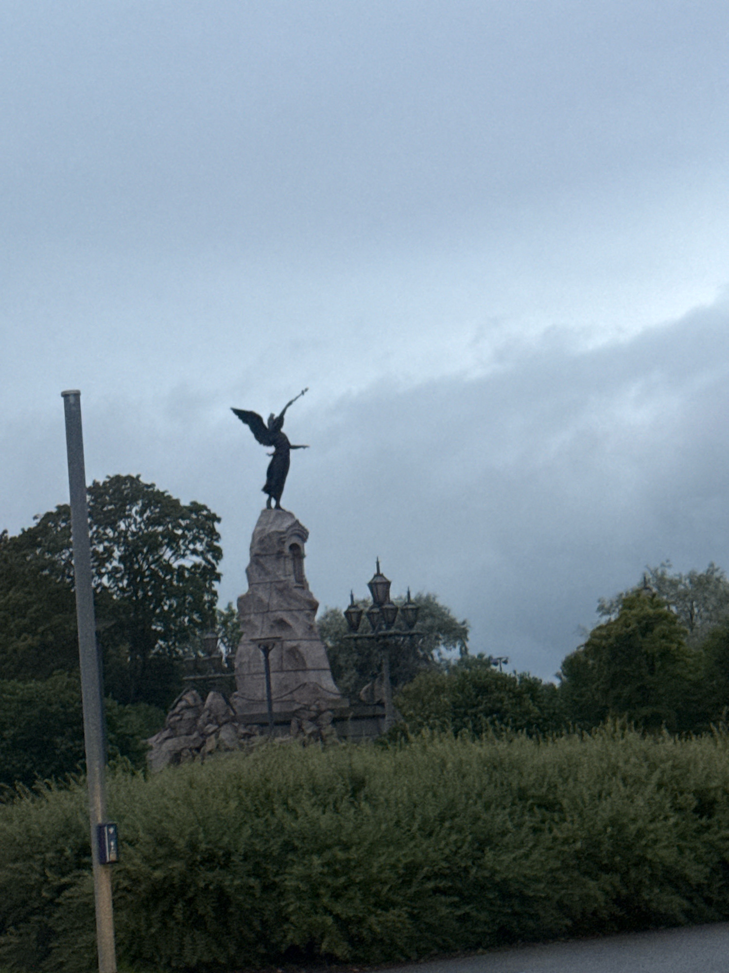 The image shows a statue of an angel on top of a stone monument. The angel appears to be made of dark metal and has its wings outstretched. The monument is made of rough-hewn stones and has a weathered appearance. In the background, there are trees and a cloudy sky. A lamppost and a signpost are visible to the left of the monument. The scene appears to be a memorial or a public monument in an outdoor setting. The image is taken from a slightly low angle, looking up at the statue. The overall mood of the image is somber, possibly due to the overcast weather and the nature of the monument. The location is not identifiable as Tallinn, Estonia based on the visual elements in the image.