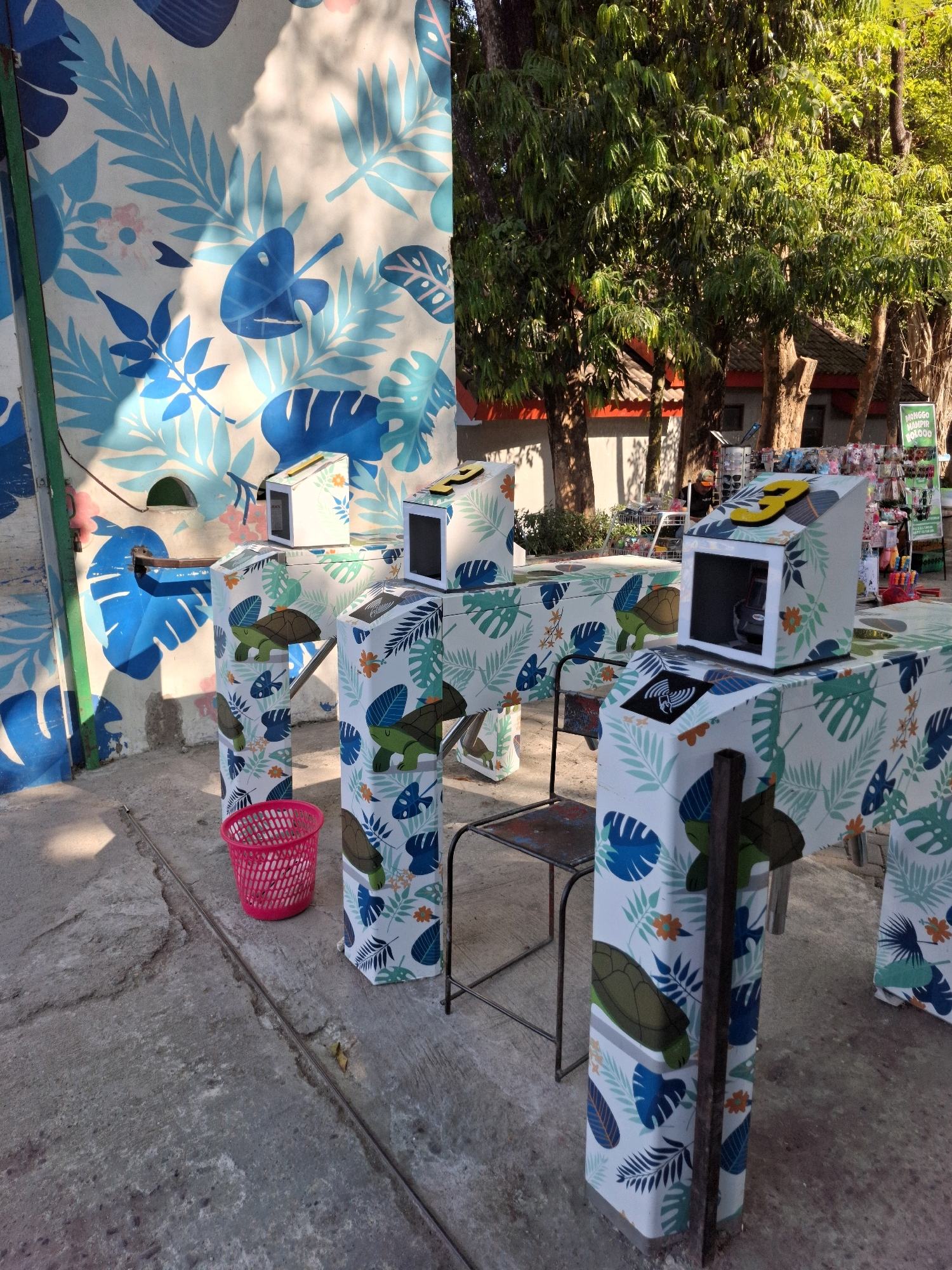 The image shows an outdoor scene in front of a building with a mural on the wall. The mural features a blue and white tropical leaf pattern. In the foreground, there are three ticket-scanning turnstiles, decorated with a similar tropical leaf and turtle pattern. A pink plastic trash can is next to the leftmost turnstile. Behind the turnstiles, a metal chair is visible. In the background, trees with green foliage are prominent, and a glimpse of a building with a red roof can be seen. To the far right, a rack of small items, possibly souvenirs, is displayed. The ground is paved with concrete. The scene appears to be at an entrance to an attraction or venue in Ngawi, Indonesia, suggested by a partially visible sign in the background that reads "MONGGO KAMPIR BOLOD."