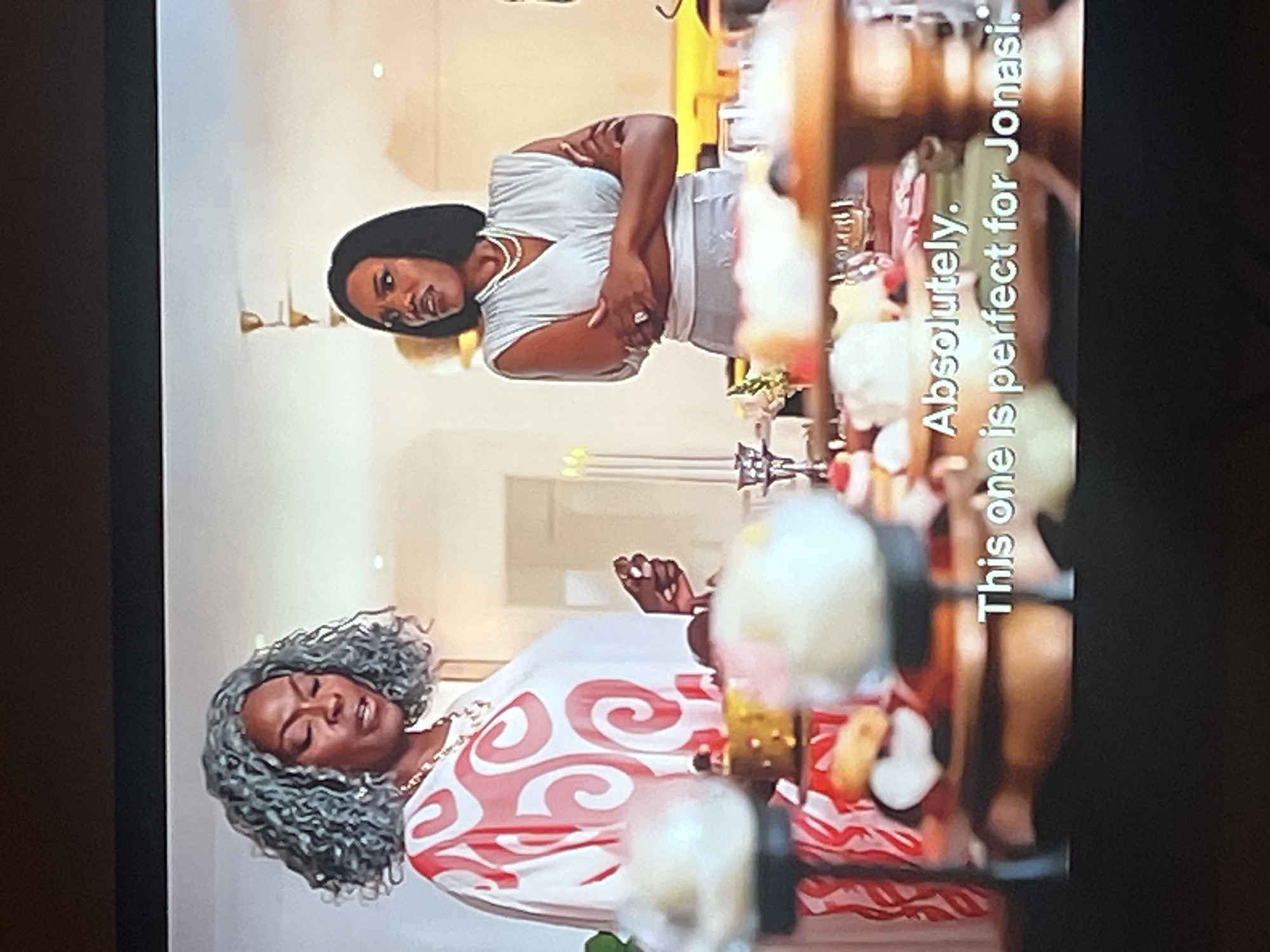 The image shows two Black women inside what appears to be a brightly lit room. The woman on the left has dark, short hair and is wearing a white V-neck top. Her arms are crossed and she is looking to her right with a neutral expression. The woman on the right has gray, curly hair and is wearing a white top with a red swirl pattern. Her eyes are closed, and her head is tilted back, suggesting a moment of reflection or emotion. She is wearing a gold necklace.
In the background, on the right side of the image, there is a blurred display of food items, possibly desserts or sweets, arranged on shelves. Some of these items are white, red, and yellow. There is also visible text overlaid on the image. From top to bottom, the text reads: "Absolutely." and "This one is perfect for Jonasi." The lighting suggests it is daytime, and there are no indications of weather or specific location cues within the room itself, other than the overlaid text which may imply a context related to Nigeria, given the provided location context.