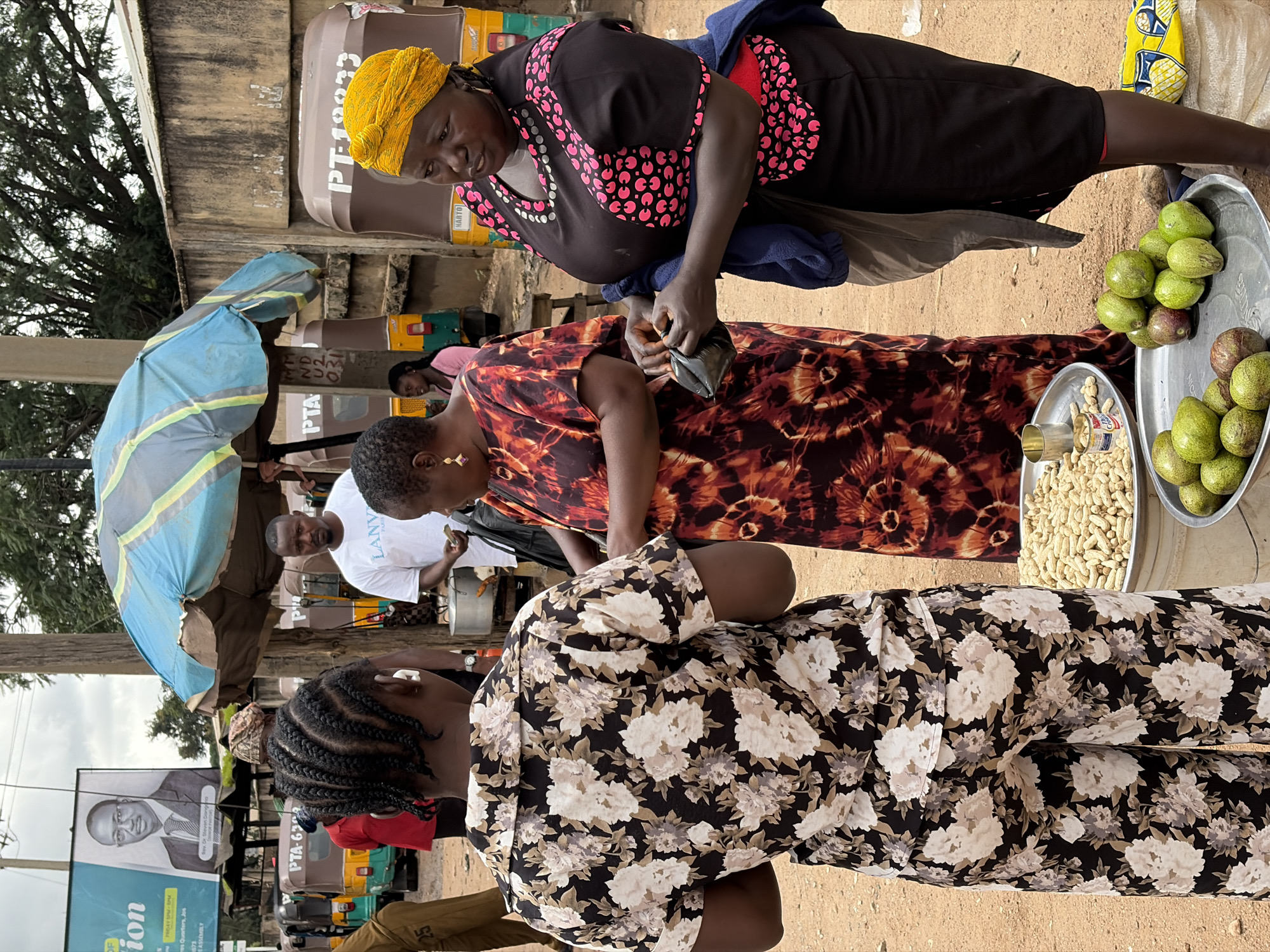 In Jos, Nigeria, a street scene unfolds with three women engaged in what appears to be a market transaction. In the foreground, a woman wearing a black floral print jumpsuit is facing away from the camera, her back to the viewer. To her right, a woman in a red and orange tie-dye dress is holding a small black item, possibly a purse or wallet, with her hands. She is looking down and appears to be interacting with the woman to her right. The third woman, on the far right, is wearing a black top with pink circular patterns and a yellow headscarf. She is looking towards the woman in the floral print and seems to be holding a small black wallet, also possibly engaged in a transaction.
In the background, other individuals are visible. A man in a white t-shirt stands near a food vendor's stall, which has a pot on a stand. Several auto-rickshaws, commonly known as 'keke napep' in Nigeria, are parked behind the people, suggesting a busy transportation hub or a roadside market. One of these vehicles has the registration "PTA-6302". A large blue and green umbrella provides shade over some of the market stalls. A billboard featuring a man in a suit and the text "OF Election" and "Dr. Steven Orovwaghere" is prominently displayed on the left side of the image, indicating a political context. The ground is sandy and dusty, typical of many Nigerian urban environments. The overall scene depicts a vibrant, everyday street life with elements of commerce and local culture.
