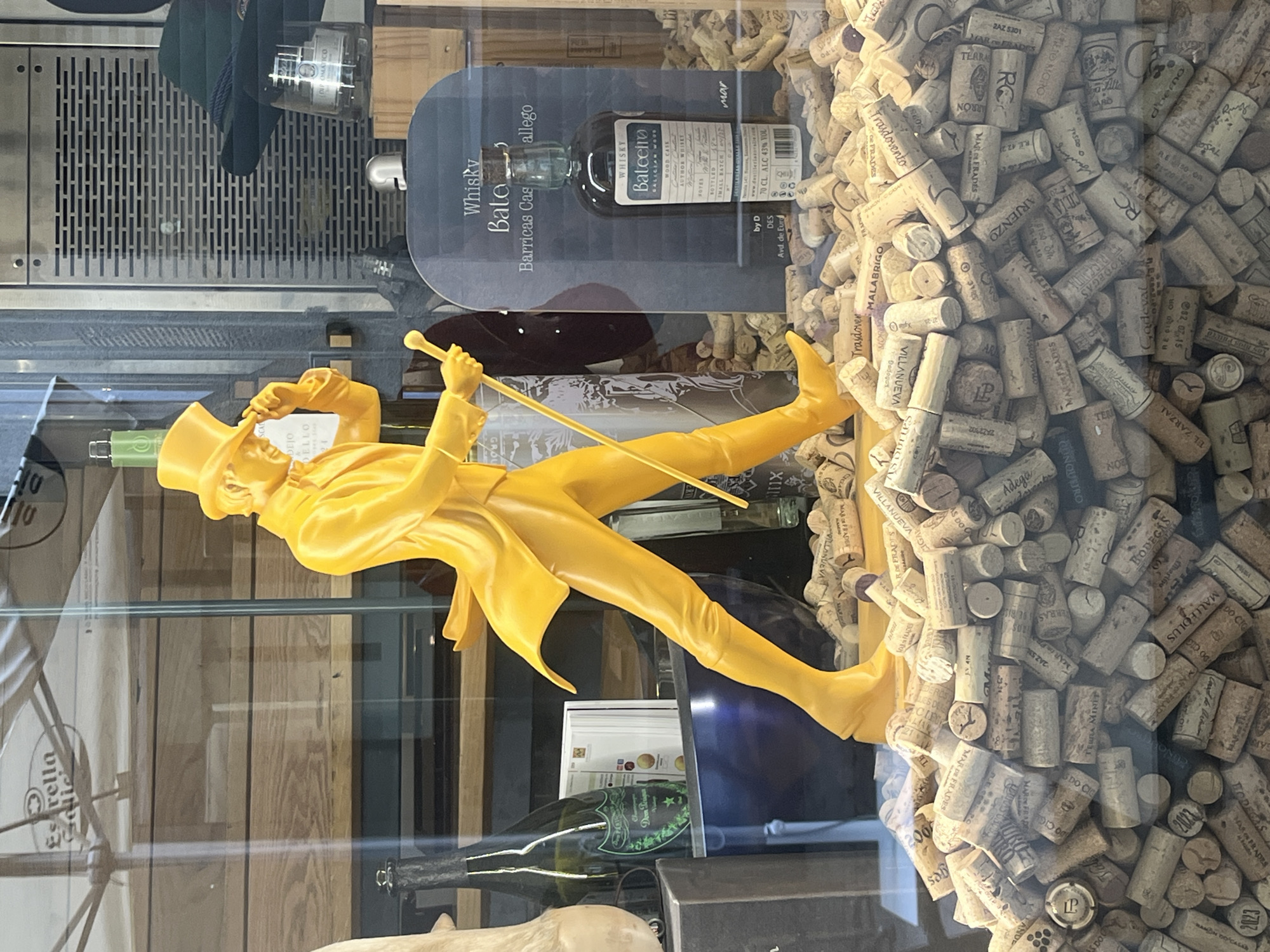 The image depicts a window display in A Coruña, Spain. The display features a gold-colored figurine of a man in a suit and top hat, leaning on a cane. Behind the figurine is a selection of liquor bottles, including a bottle of "Whisky Balteiro" and a bottle of "Dom Perignon" champagne. The background is filled with used wine corks. The location is implied by the "Balteiro" label, referencing a Galician distillery, as well as labels on corks such as "Mar de Frades".