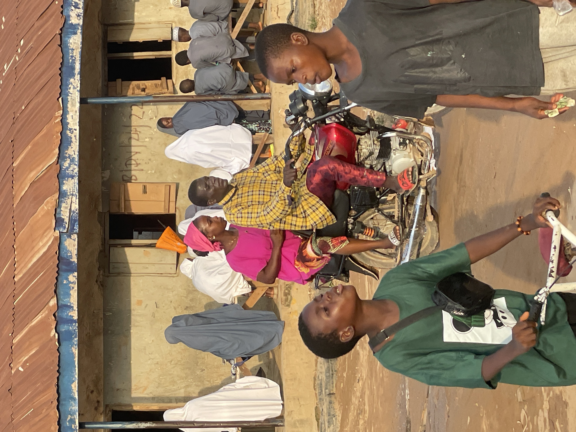 In the city of Abuja, Nigeria, a group of people are gathered outdoors in front of a building with a corrugated metal roof. A man and woman are on a red motorcycle, with the man driving and the woman sitting behind him. The man is wearing a yellow and black plaid shirt and red patterned pants. The woman is wearing a pink headscarf and a pink shirt with a colorful skirt. Two young boys are in the foreground, one in a green t-shirt with a Mickey Mouse graphic and the other in a dark t-shirt. Behind them, several people are sitting on a bench, many of them wearing white or gray robes. The building has a concrete facade, windows with wooden shutters, and a blue trim beneath the roof. Graffiti is visible on the building's wall. The ground is dirt.
