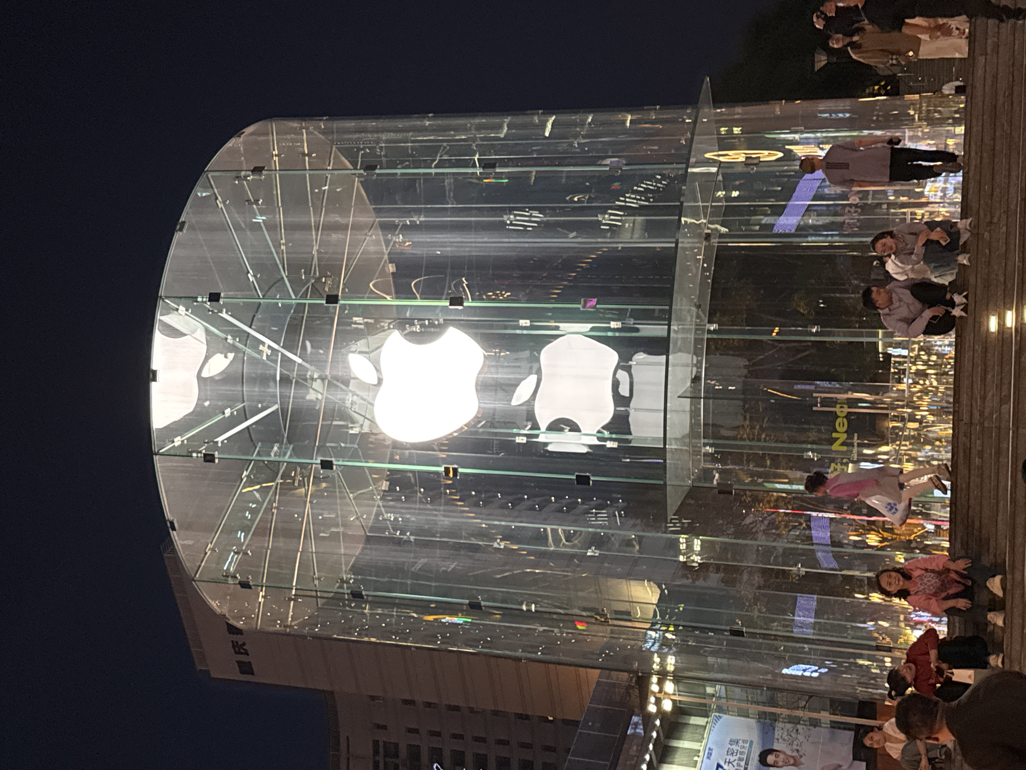 The image shows an Apple store in Yuzhong District, China at night. The store has a cylindrical glass structure with the Apple logo prominently displayed. People are standing and sitting on the steps next to the store. Buildings are visible in the background.