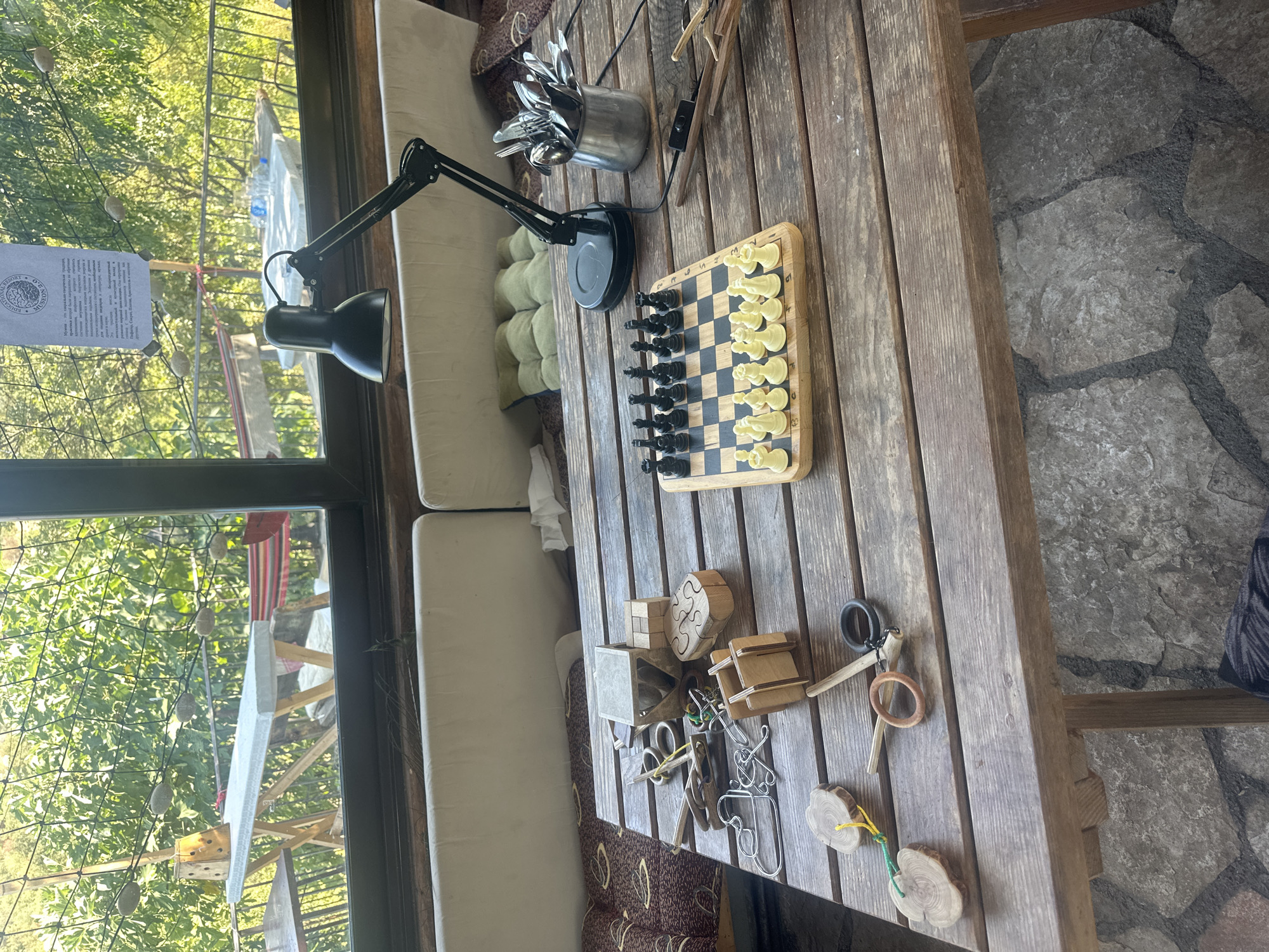 The image shows a wooden table with various objects on it, including a chessboard set up for a game, several wooden puzzles, metal puzzles, two wooden rings, and a metal container filled with cutlery. A black desk lamp is positioned behind the chessboard. The table is situated on a patio or balcony overlooking a lush green outdoor area with trees and foliage visible through a window. In the background, there are outdoor tables and chairs, as well as a white paper with text hanging from a string. The flooring beneath the table is made of large, irregular stones. The overall scene suggests a relaxed, possibly recreational or educational setting. While the image is taken in Cetinje, Montenegro, the specific location within the city is not identifiable from the visual information provided. There are no people visible in the image.