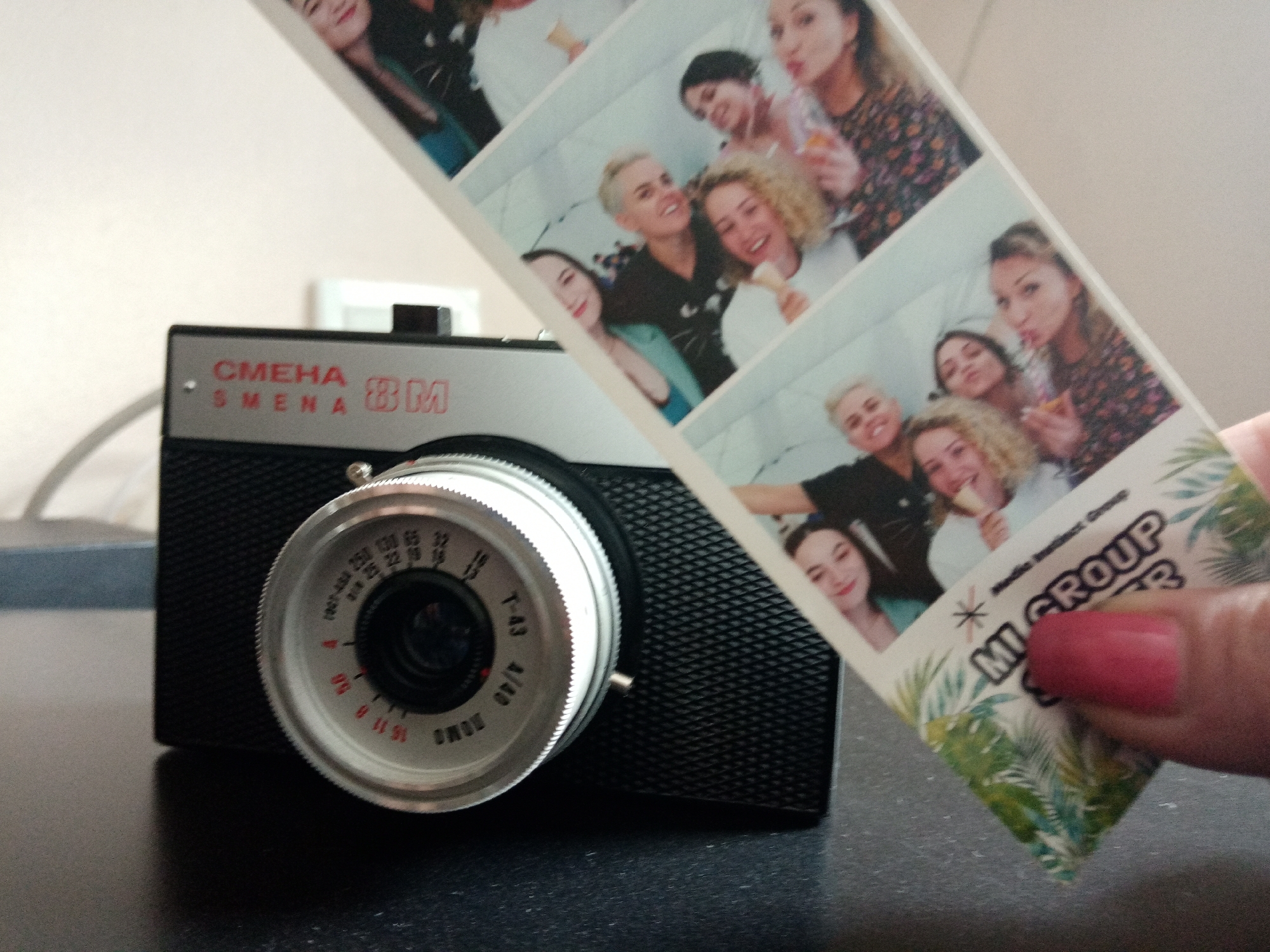 The image displays a vintage Smena 8M film camera positioned on a dark, textured surface, likely a table or countertop. The camera's top plate features "СМЕНА" (Smena in Cyrillic) and "SMENA 8M" in red lettering. Its prominent silver lens housing is marked with "T-43 4/40" and "LOMO," along with various settings for film speed (e.g., "ASA 250," "ASA 125," "ASA 65," "ASA 32," "ASA 16," "ASA 8") and aperture values ("16 11 8 5.6 4").
A person's hand, with pink nail polish, holds a photobooth strip diagonally across the upper right portion of the frame. The strip contains multiple individual photographs of young women, all appearing happy and engaged. Some are smiling broadly, while others make playful expressions. They are seen holding what resembles ice cream cones or drinks, suggesting a fun, social event or gathering. Visible text on the photobooth strip includes "MI GROUP" and a smaller "Studio Instinct Group." The background behind the camera is a plain, light-colored wall. The presence of the Soviet-era Smena camera, set against the backdrop of a modern photobooth strip depicting cheerful friends, juxtaposes classic photography equipment with contemporary social imagery. The camera model is consistent with the location context of Elektrostal, Russia.