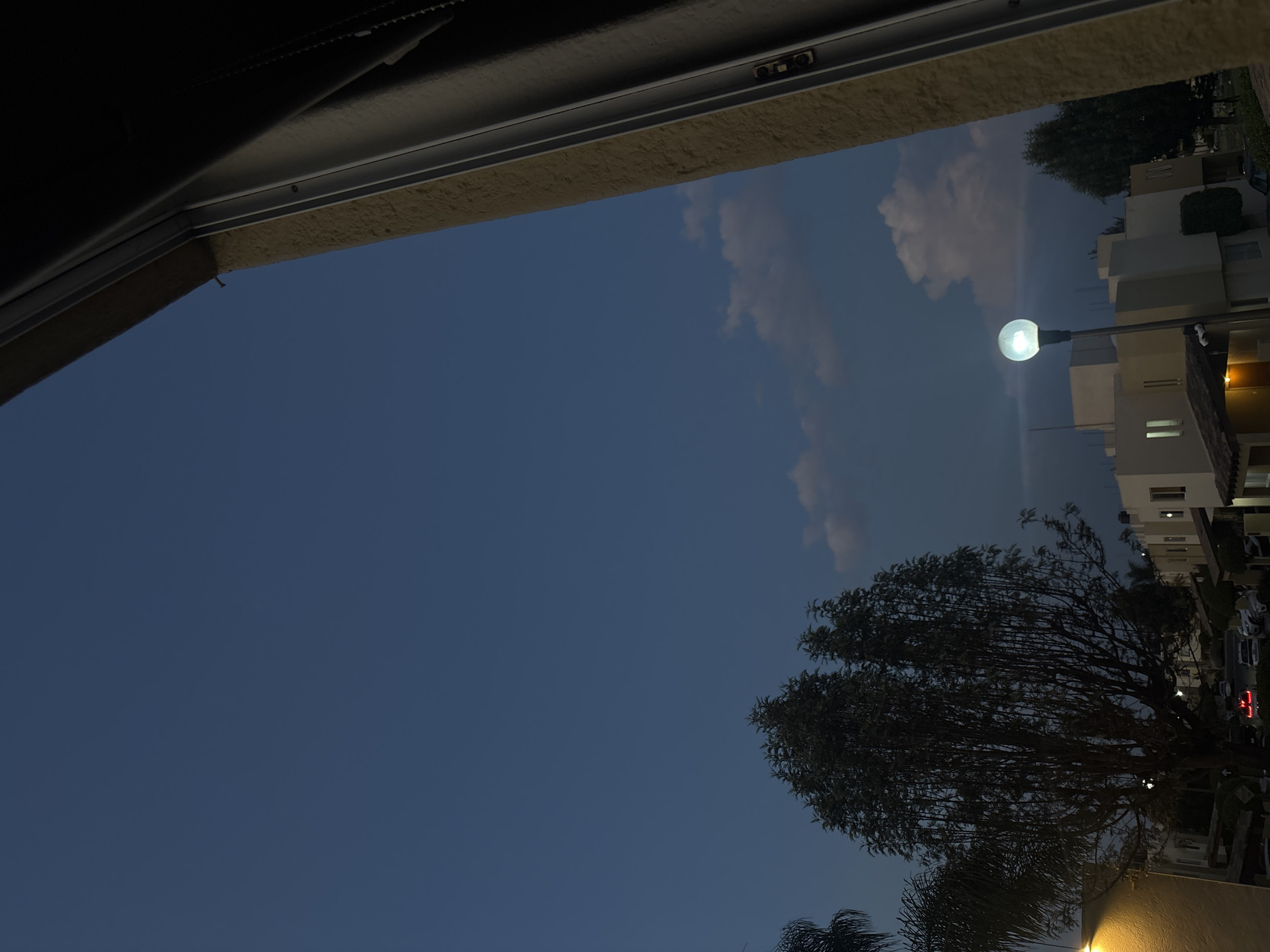 A low-angle, indoor view looks out through a window or opening in Puebla, Mexico. The exterior features a darkening blue sky with some white clouds. A street light is illuminated near a row of several-story buildings, which appear to be apartment complexes, illuminated by some internal lights. A large tree is in the foreground, silhouetted against the sky and buildings.