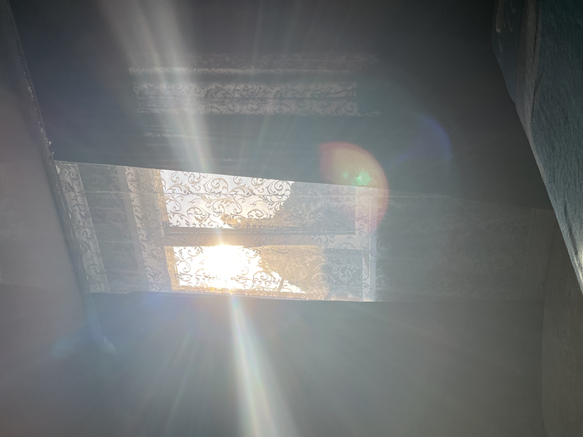 This is a medium shot, eye-level interior photograph looking towards a window with sunlight streaming through, creating lens flares and a hazy atmosphere. The window is partially covered by decorative sheer curtains with a damask pattern and dark, opaque curtains on the right. Outside the window, foliage is visible, silhouetted against the bright sun. The room appears to be a bedroom, with a bed covered with a blue patterned comforter in the lower right foreground. The walls are dark, contrasting with the bright light entering the room. The image was captured in Saratov, Russia.