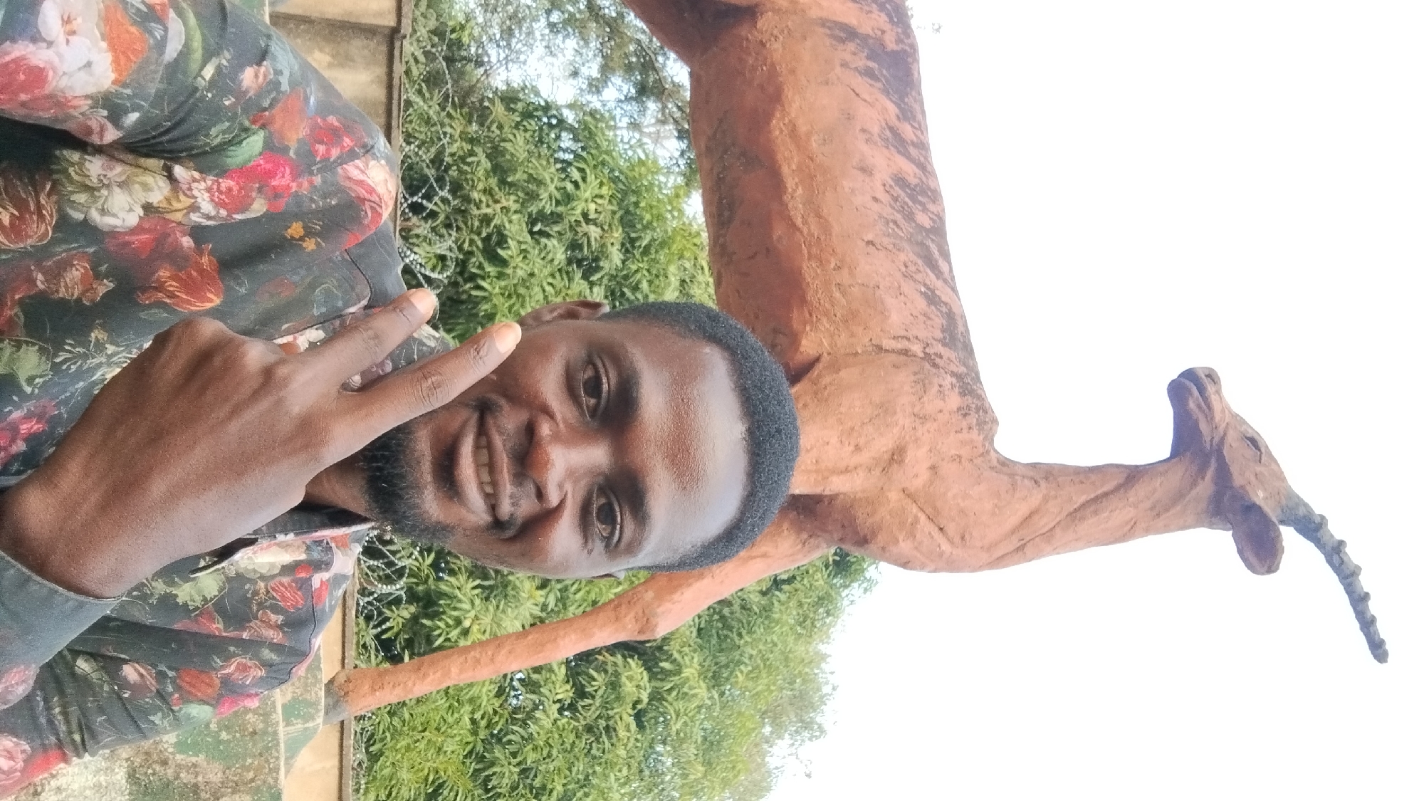 The image is a selfie of a smiling man giving a peace sign, taken outdoors in front of a statue of an antelope. The man has short black hair, a dark complexion, and a short beard. He wears a dark shirt with a colorful floral pattern.
Behind him and slightly to the right is a reddish-brown statue of an antelope, possibly constructed from concrete or a similar material. The statue appears to be standing, with its head turned slightly. The sky is visible in the background. There are green trees and possibly other foliage visible. Barbed wire is present near the man. The photo appears to have been taken in Jos, Nigeria.