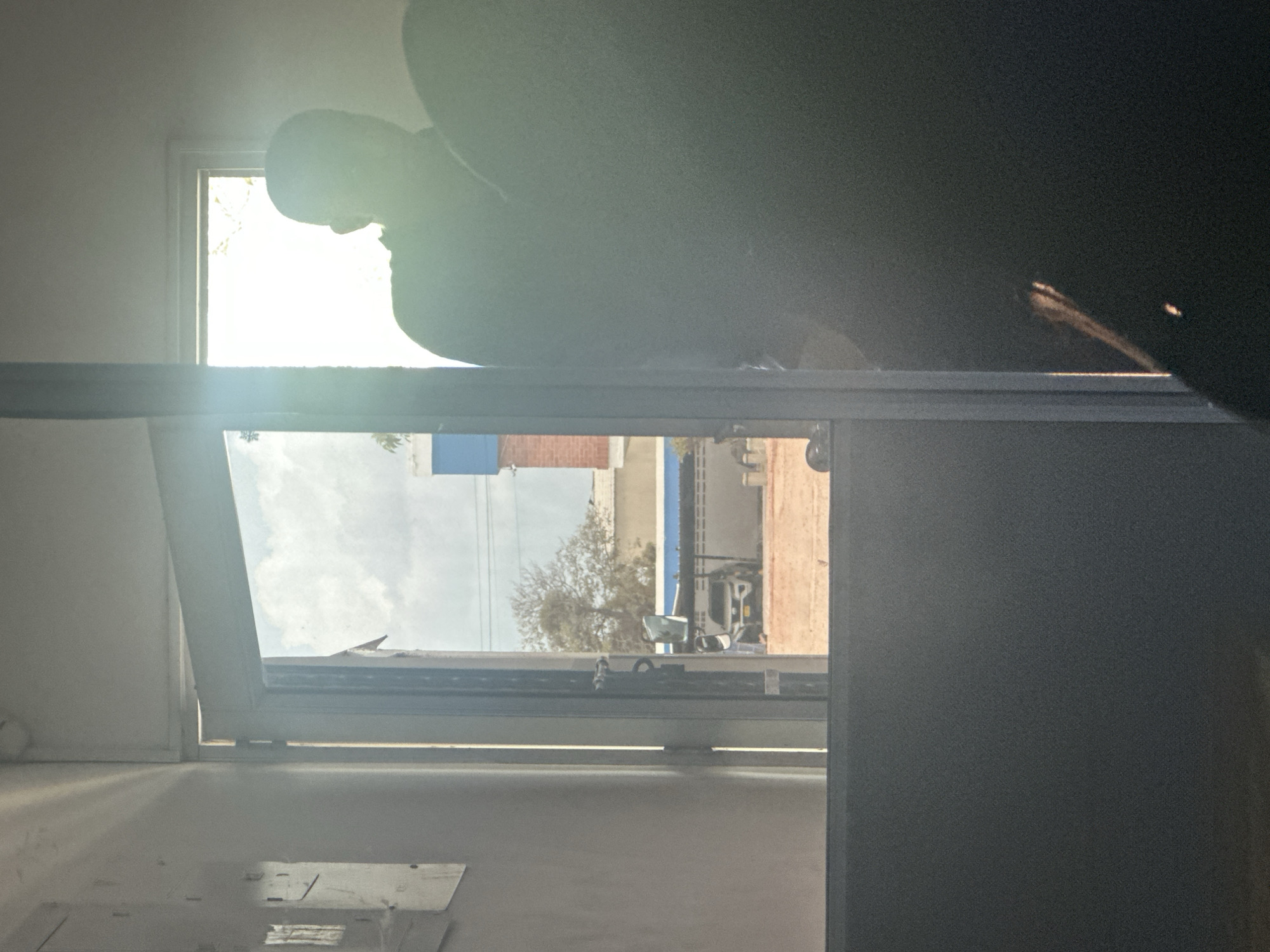 The image captures a view through an open doorway, looking outwards into an urban environment. The foreground is dominated by a dark, out-of-focus mass, possibly a person or furniture, obscuring the lower right portion of the scene. The doorway itself is framed by a light-colored wall on the left and a bright, vertical metal beam on the right. Sunlight streams in from the right, creating a strong glare and silhouetting a person's head and shoulders visible in the middle ground, standing just beyond the doorway. This person appears to be wearing dark clothing.
Through the doorway, the exterior scene shows a paved area with a white SUV parked under a covered structure. Beyond the car, there is a low building with a white wall and a blue trim. Further in the background, a bare-branched tree is visible against a bright, cloudy sky. The overall impression is a moment captured in daylight, with significant contrast between the brightly lit exterior and the darker interior. The scene is set in the city of Juan Mina, Colombia.