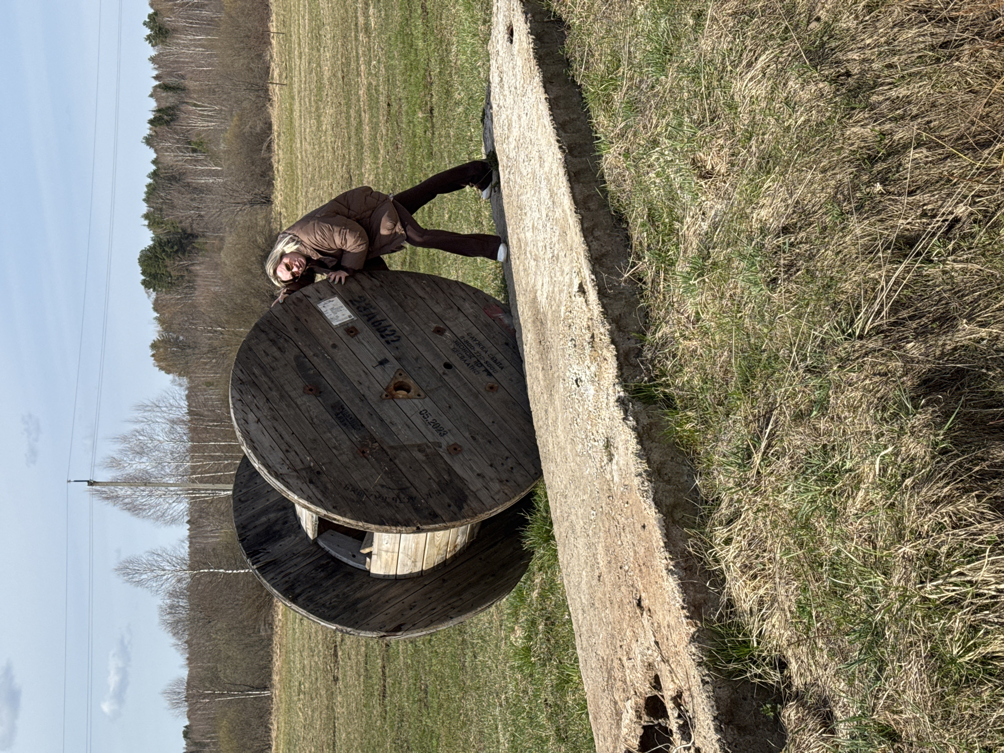 A woman in a brown jacket and pants is leaning on a large wooden cable reel and a concrete barrier in a grassy field. Trees are visible in the background, and power lines can be seen in the sky. Based on the information provided, the location is Уральское, Russia.