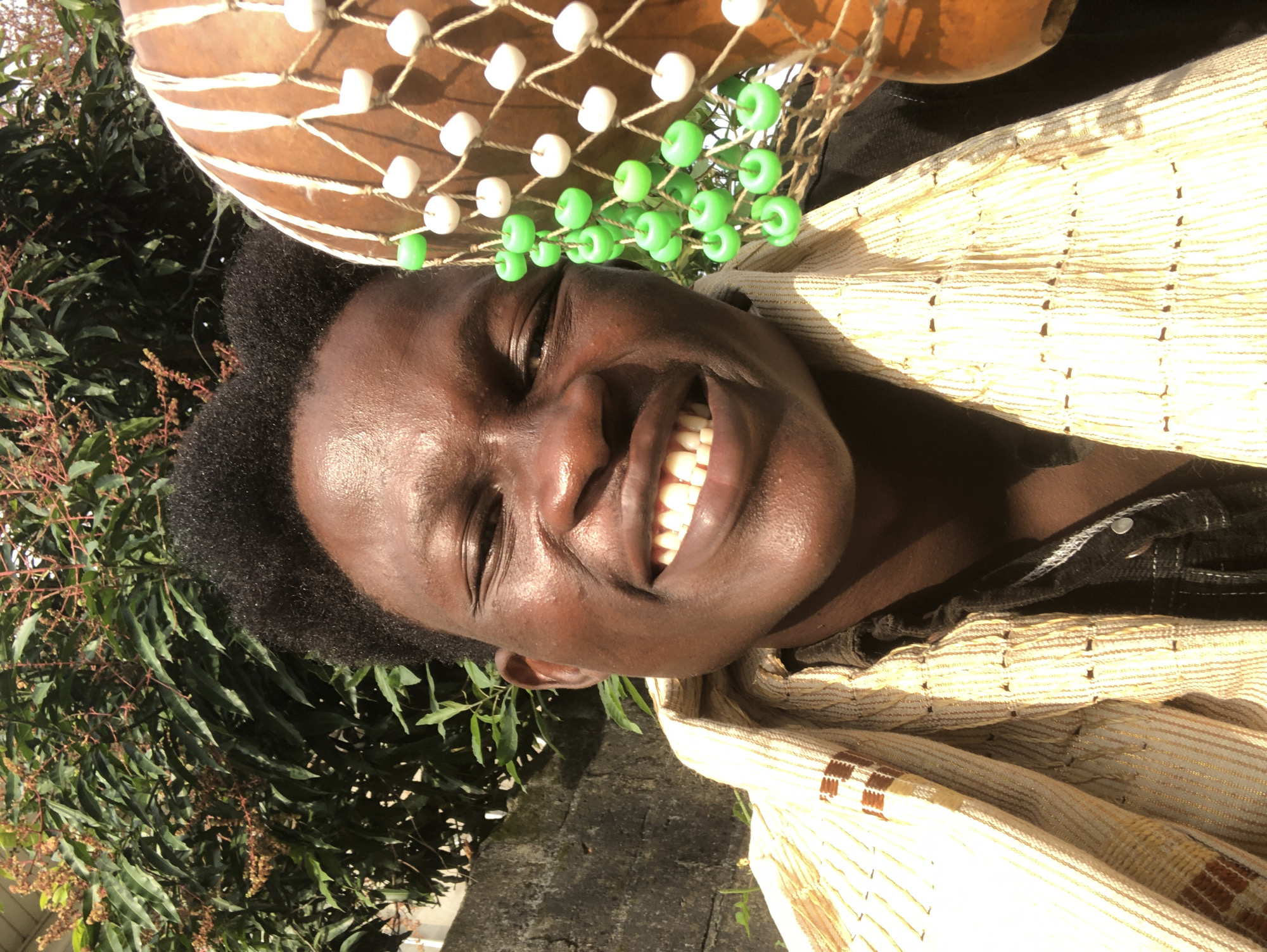 A close-up, outdoor, selfie-style portrait features a young Black male in Ado, Nigeria. He is smiling widely, exposing his teeth. His dark hair is short and styled in an afro. He is wearing a light-colored garment with a striped pattern, draped around his neck and shoulders, over a dark shirt. To the right of the frame, a portion of a stringed gourd instrument is visible, adorned with white and green beads. The background consists of lush green foliage, suggesting a tropical environment, with a hint of a stone wall on the left side. The lighting indicates it is daytime, with the sun casting shadows and highlighting the subject's features.