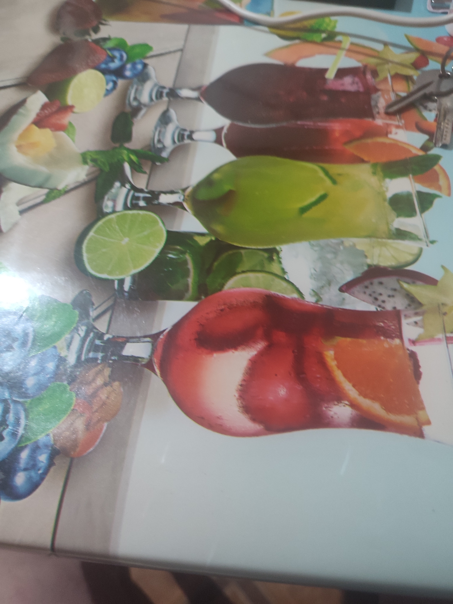 The image depicts a still life arrangement of colorful fruit-filled cocktails and garnishes on a reflective surface, possibly a table or countertop. The arrangement includes glasses filled with red and green liquids, garnished with various fruits such as oranges, limes, strawberries, blueberries, dragon fruit, and starfruit. Some glasses contain ice and straws. The background appears to be a textured surface, possibly wood grain. There are no people or identifiable landmarks of Chernivtsi, Ukraine, visible in the image.