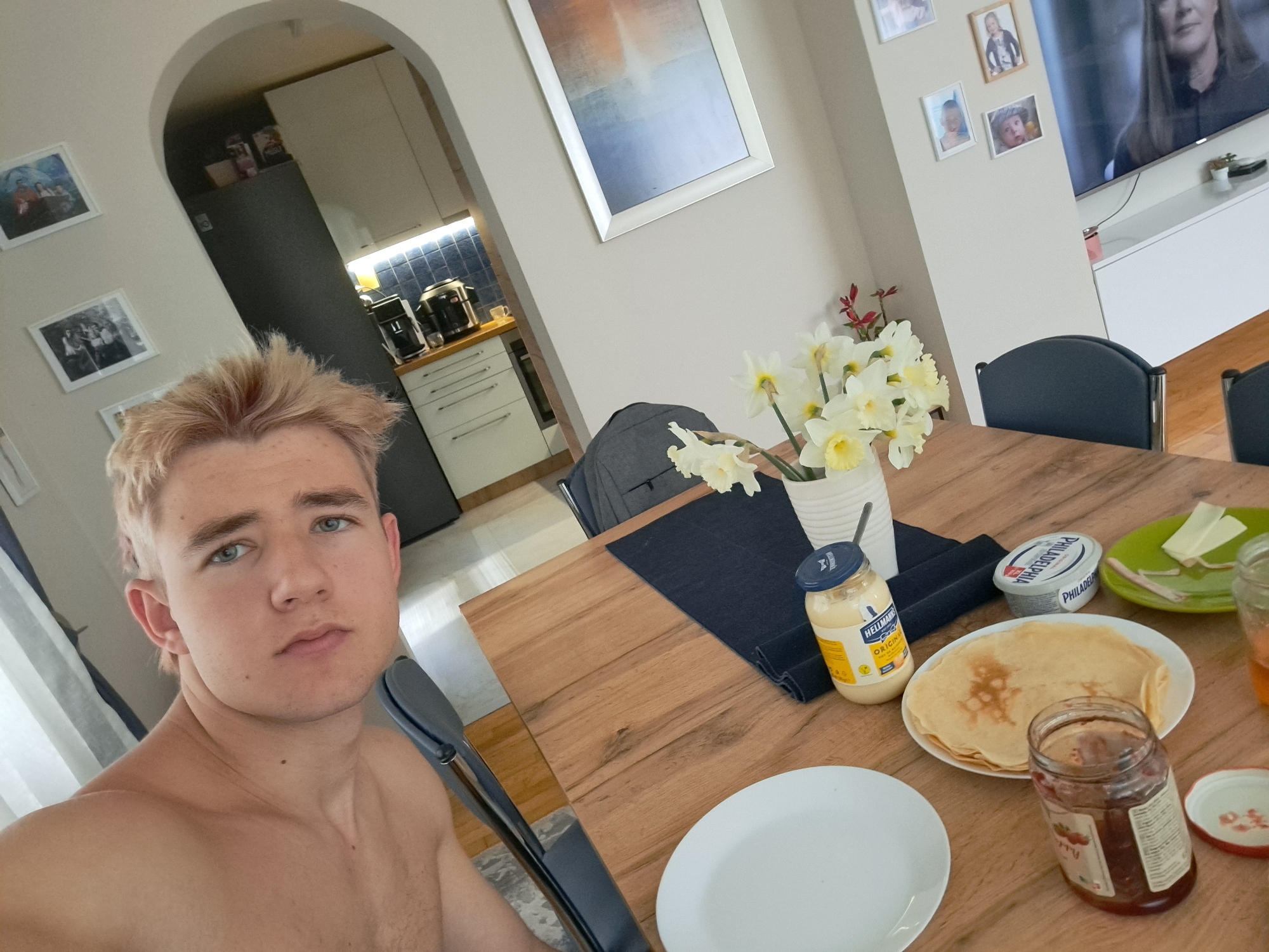 The photograph depicts a fair-skinned young man with blonde hair sitting at a wooden dining table. He appears to be taking a selfie. He is shirtless.
On the table, there is a white plate, a stack of pancakes, a jar of jam, a jar of mayonnaise, a container of cream cheese, a small plate with cheese slices, and a vase of daffodils. A dark blue cloth is under the vase and mayonnaise.
In the background, there is an arched doorway leading into a kitchen with white cabinets and stainless-steel appliances. There are framed pictures on the wall. A television is visible in the background to the right.
While the location cannot be definitively confirmed as Sofia, Bulgaria from the image alone, the style of the apartment and the presence of European products suggest a European urban environment.